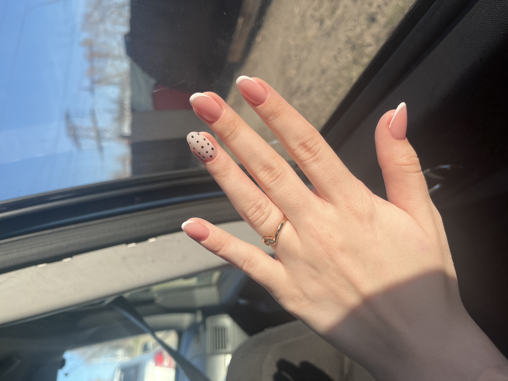 The image displays a person's right hand, showcasing a fresh manicure and a ring, set inside a vehicle in Chaikovsky, Russia. The hand is illuminated by bright sunlight, casting distinct shadows and suggesting it is daytime on a clear, sunny day.
The nails are neatly manicured, with most fingers featuring a classic French tip style: a natural pink or nude base topped with clean white tips. The ring finger's nail stands out with a playful design of small black polka dots on a light, neutral base. A golden ring, characterized by a unique swirling design, is worn on the middle finger.
The setting is an interior of a car, indicated by the visible window frame, dark interior trim, and reflections on the glass. Through the window, blurry shapes of what appear to be buildings or structures in the background are visible, consistent with an urban or suburban environment in Chaikovsky, Russia. There is no specific activity taking place other than the hand being presented for the photograph. No visible text is present in the image.