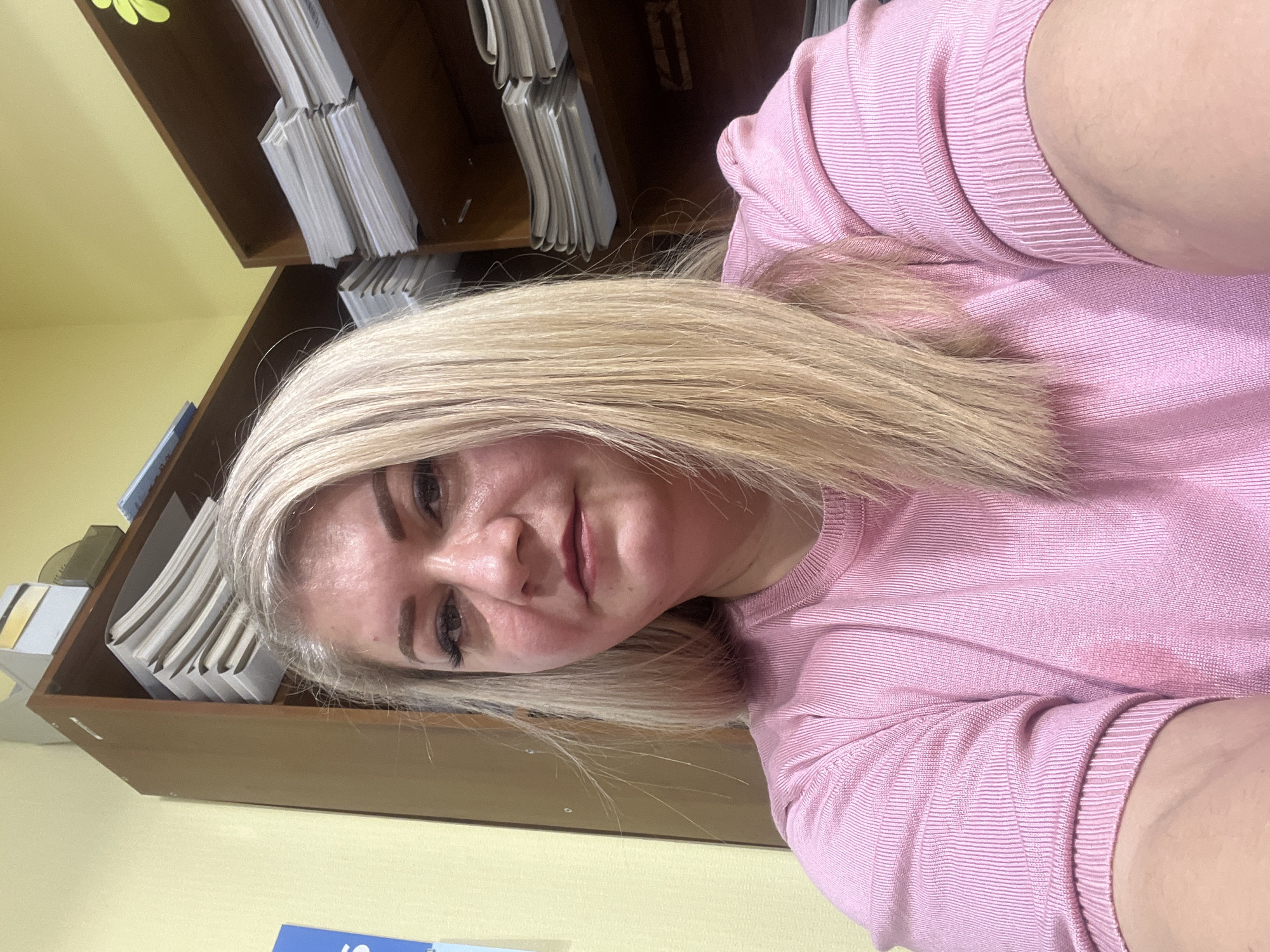 A woman with blonde hair wearing a pink shirt is positioned in front of a brown shelving unit filled with files or paperwork. The background includes a pale yellow wall and a glimpse of a blue object. The image does not contain enough information to identify a specific location in Izhevsk, Russia.