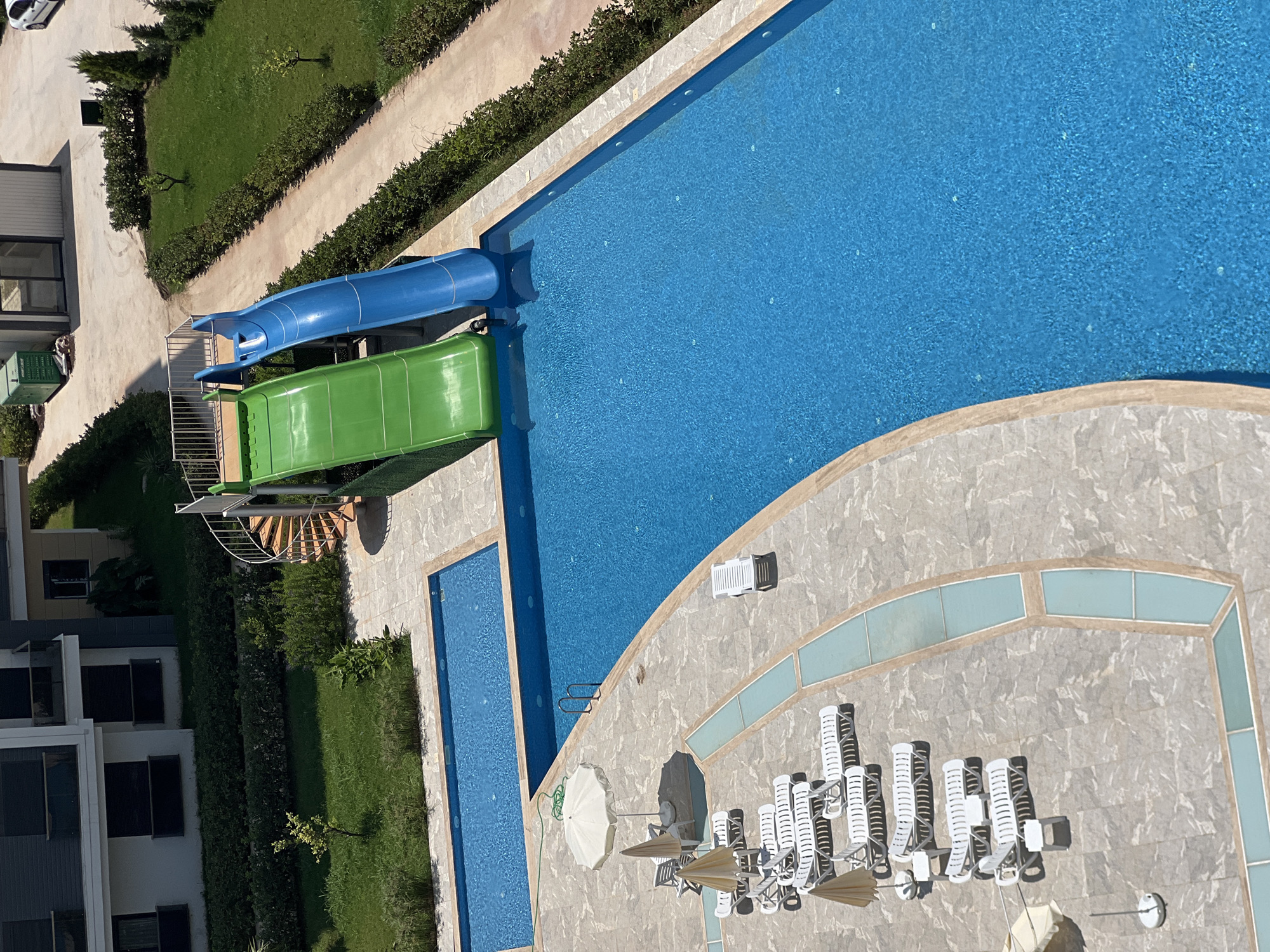 This is an aerial, eye-level, medium shot of a resort swimming pool area with two slides. The swimming pool is large and blue. To the left of the pool, there are rows of white lounge chairs with a few white and beige umbrellas. To the right of the pool is a grassy area with a hedge, and beyond that, a paved path and a building. There are no people in the image. The scene appears to be in Aksu, Turkey, a popular tourist destination known for its hotels and resorts.