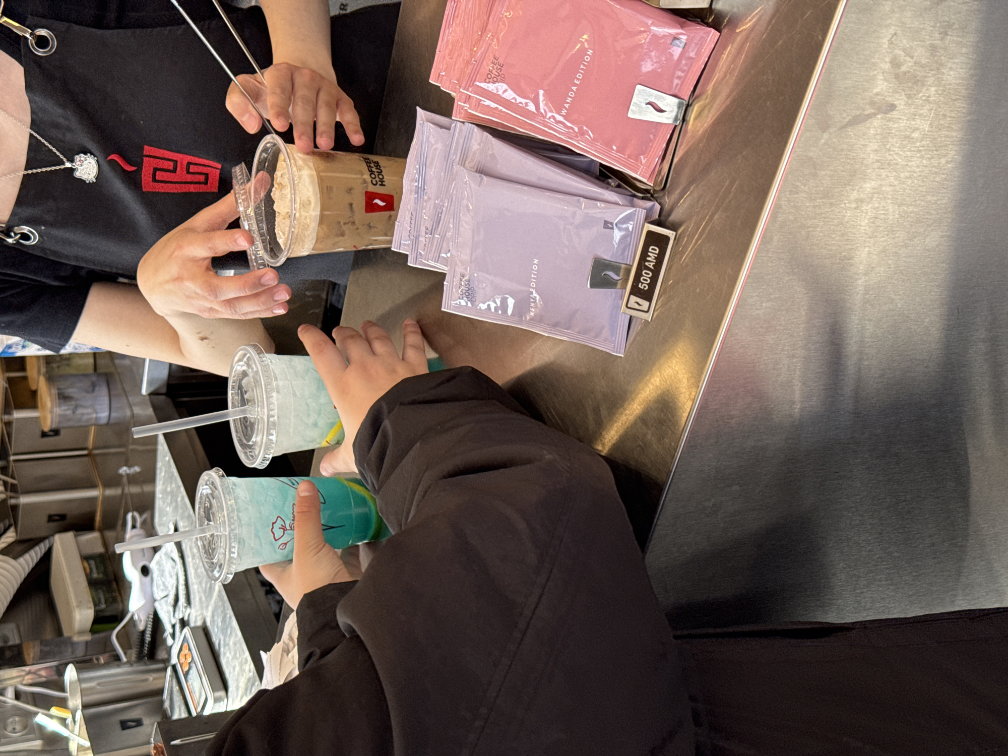 The image shows a transaction taking place at what appears to be a coffee shop in Yerevan, Armenia. A person wearing a black apron with a red logo and a silver Hello Kitty necklace hands a customer an iced coffee drink. The customer is holding two blue-tinted beverages in clear plastic cups with straws. On the counter are packets of drip coffee labeled "Kenya Edition" and "Wanda Edition", with one of the Kenya packets marked "500 AMD", suggesting the price in Armenian Dram. The counter itself appears to be made of metal, and a cash register or similar device is visible on the left side of the image.