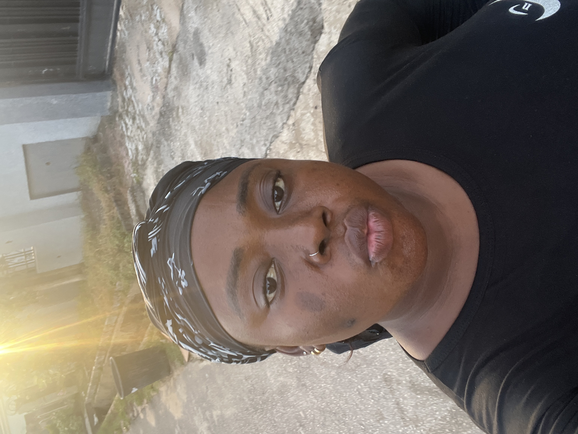 A person with dark skin and dark eyes looks directly at the camera, wearing a black patterned headscarf and a black long-sleeved top. A small ring is visible in their left nostril, and they wear earrings. The background appears to be an outdoor area, possibly a street or sidewalk. There is a building to the left, and sunlight appears to be shining from the left side of the frame. The specific location within Abuja, Nigeria is not discernible from the provided image.