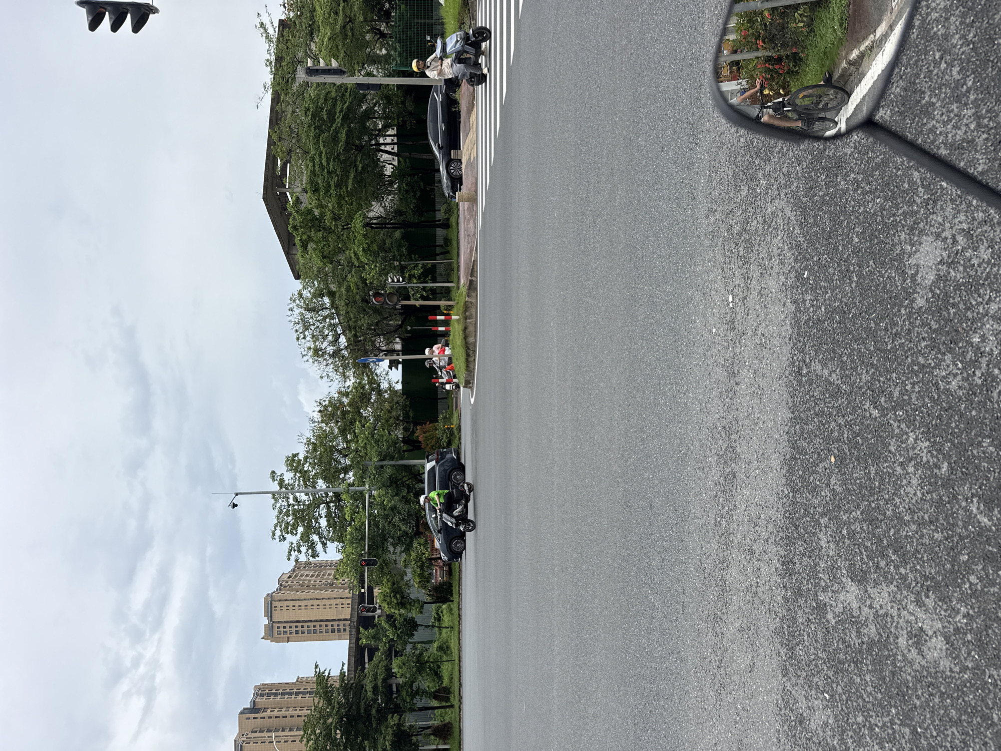 The image captures a street scene in Huizhou, China, featuring an intersection with vehicular traffic. In the foreground, the asphalt road is visible, with a motorcycle side mirror reflecting a cyclist. The main part of the scene shows several vehicles at the intersection. A person in a green jacket rides a black electric scooter, heading towards the right of the frame. A dark sedan is parked behind the scooter. Further to the right, another person on a similar electric scooter, wearing a yellow helmet and white top, is crossing a pedestrian zebra crossing. A black sedan is visible behind this scooter. In the background, tall residential buildings and trees line the street. Traffic lights are present, with some showing red signals. The sky is overcast, suggesting cloudy weather.