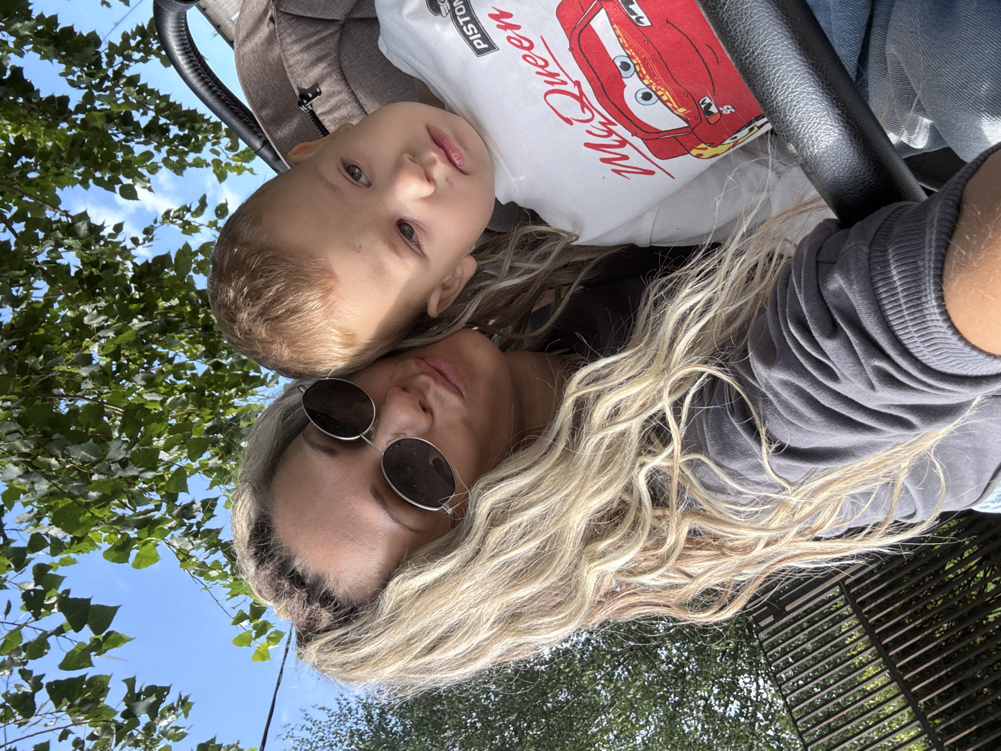 In Rusanovo, Russia, a close-up selfie is taken outdoors, showing a woman and a young child. The woman, with blonde wavy hair and sunglasses, is leaning in, pouting her lips. The child, in a stroller, wears a white t-shirt with a "McQueen" graphic. Trees and a blue sky are visible in the background. A black stroller handle is in the foreground.