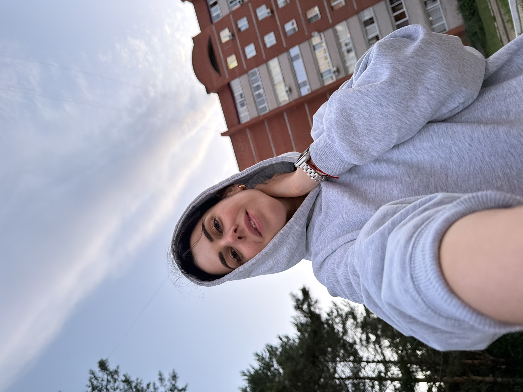 This is a selfie taken by a woman with dark hair and makeup, wearing a grey hooded sweatshirt. She is looking at the camera with a slight smile. The photo is taken from a low angle, looking up towards the sky. In the background, a tall, reddish-brown building with many windows is visible, suggesting an urban or suburban setting. There are also some trees in the foreground, out of focus. The sky is cloudy, indicating it might be overcast or late in the day. The overall mood of the image is casual and relaxed. There is no visible text in the image.