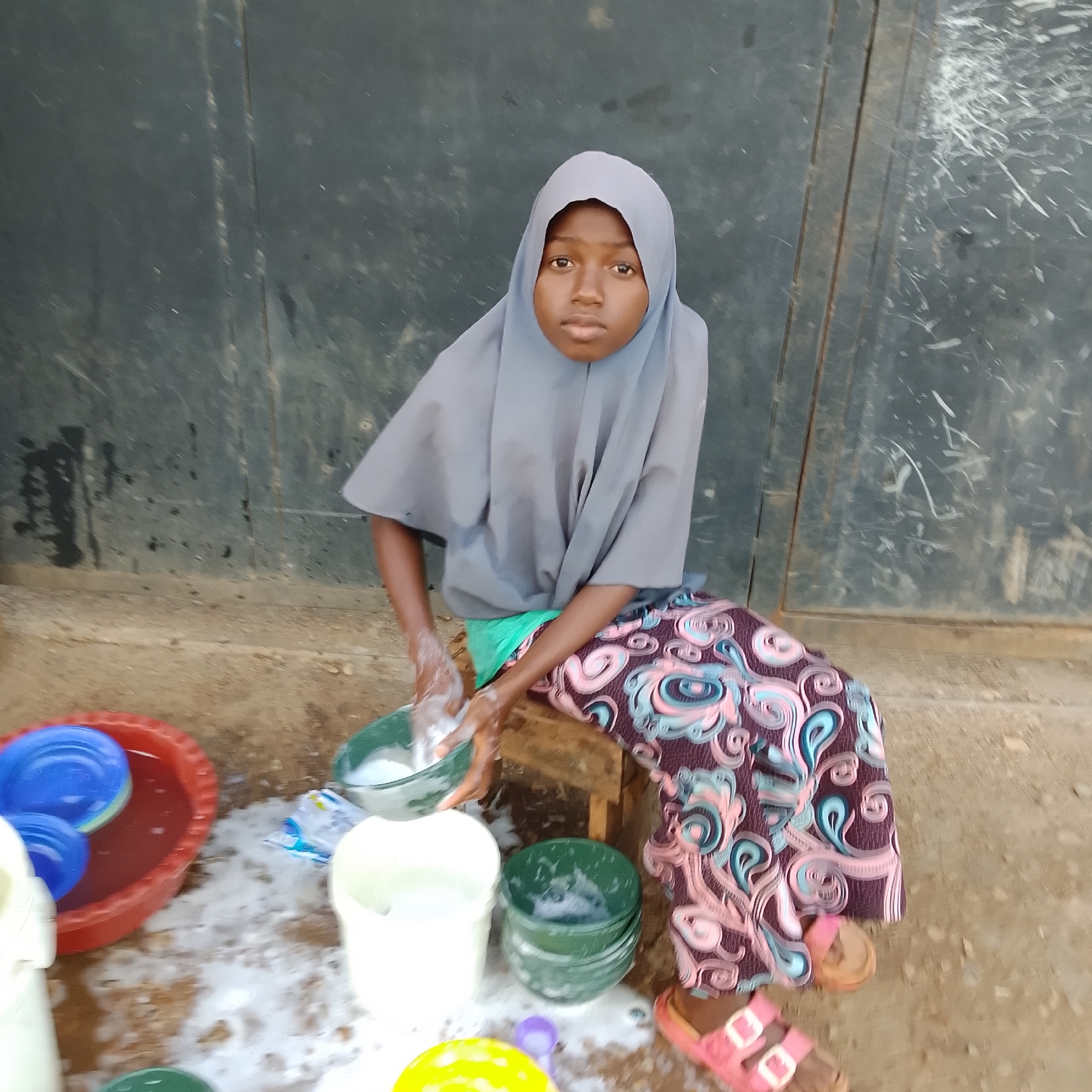 In an outdoor setting, a young girl wearing a grey hijab and a patterned skirt is seated on a low wooden stool, washing dishes. She is facing forward, looking directly at the camera with a neutral expression. Her hands, covered in soap suds, are actively engaged in washing a green bowl. To her left, a red basin holds two blue plates submerged in water. Several white and green buckets, some filled with soapy water, are arranged around her on the ground, along with scattered dish soap packaging. The background consists of a dark, weathered metal door. The ground is a mix of dirt and sand. The overall impression is one of domestic chores being performed outdoors.