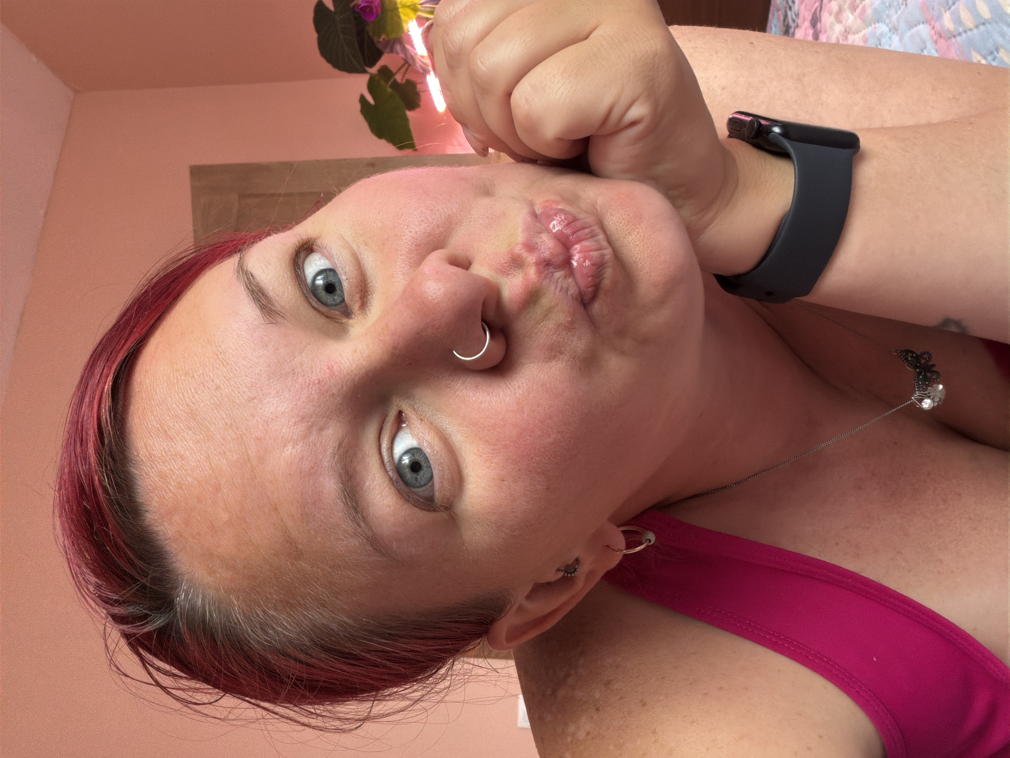 A close-up selfie captured indoors in Chaikovsky, Russia, features a person with striking blue eyes, dark red hair, and fair skin with visible freckles. The subject's lips are pursed in a playful pout, and their gaze is directed straight at the camera. A silver hoop nose ring is prominent on their face, and small earrings are visible. Their right hand is raised, with fingers curled, resting near their chin.
The person is wearing a dark pink or magenta top and a black smartwatch with a dark face on their left wrist. A delicate silver necklace with a decorative pendant, possibly a butterfly, hangs around their neck. Distinct lighter, irregular patches of skin are visible on their arm and shoulder, which could suggest vitiligo. The hair shows reddish dye with some natural lighter roots near the scalp.
The background consists of a light pink wall and part of a plant with large green leaves and a hint of purple, possibly from flowers or reflected light. A wooden surface is also partially visible. The natural, bright lighting indicates it is daytime. No visible text is present in the image.