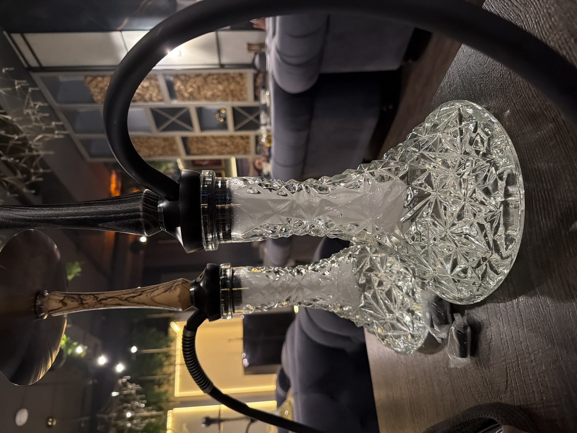 The image shows two hookahs with ornate, crystal-like glass bases, placed on a dark wooden table. The hookahs have black hoses and stems, one with a brown and black wood handle. The bases appear to be filled with a white vapor. In the background, there is a blurred view of a restaurant or lounge setting, with cushioned seating and decorative wall panels. The location might be in the city of Krasnoyarsk, Russia.