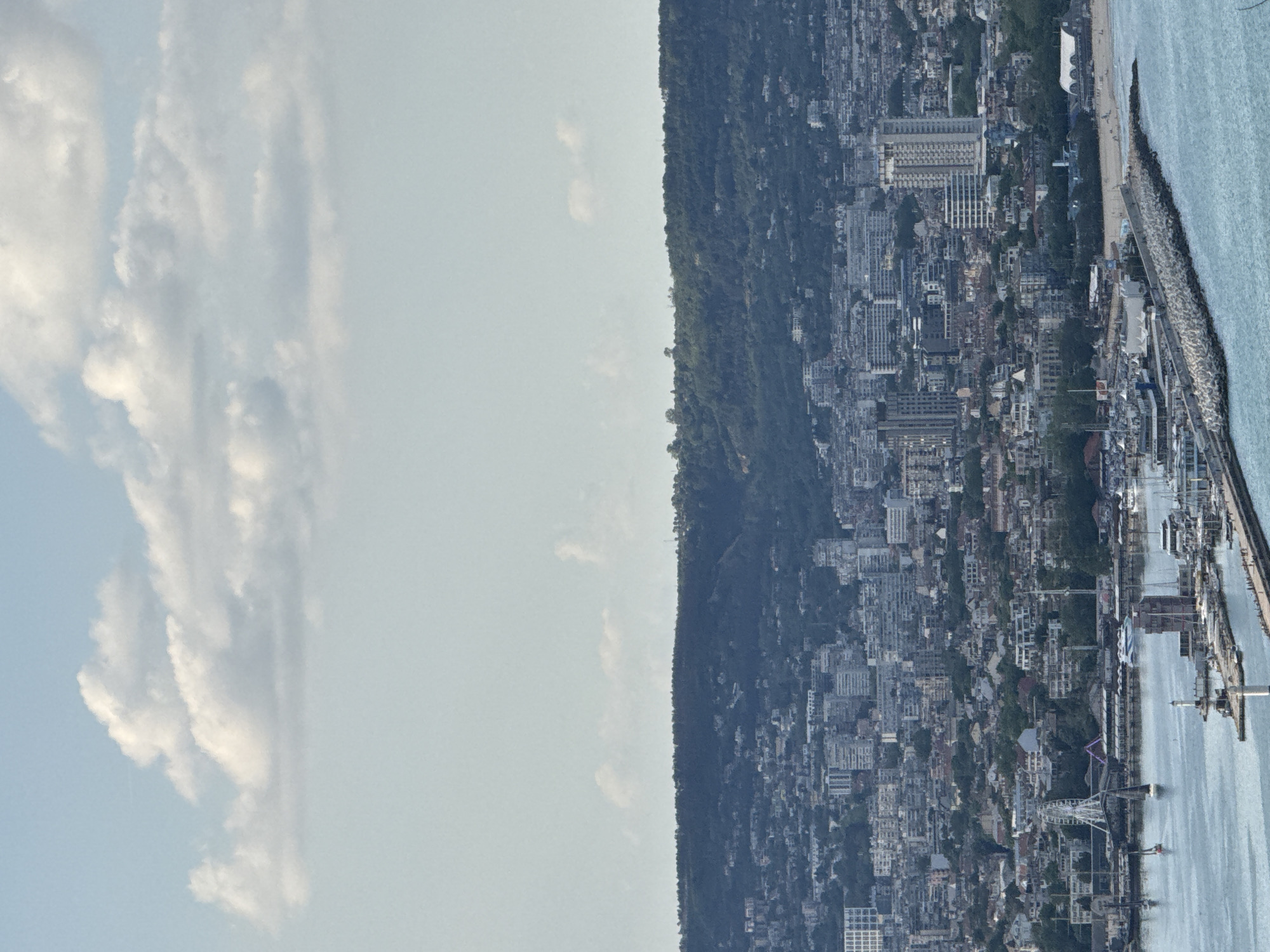 The image shows a panorama of Varna, Bulgaria, with the sea to the right and a hillside to the left. The city has a dense urban landscape, with numerous buildings of various heights. The seaside area has a long pier and some marina activity. In the distance, a mountain is visible. The sky is lightly cloudy.