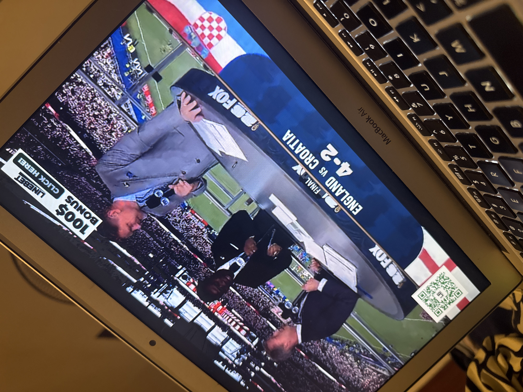 The image shows a laptop screen displaying a broadcast of a soccer match. The screen is angled, and the broadcast appears to be from Fox Sports. Text on the screen reads "FINAL ENGLAND VS CROATIA 4-2". Two men in suits are visible on the broadcast, appearing to be commentators or analysts. A QR code is also visible on the lower left corner of the screen, superimposed on a flag that appears to be the flag of England. The laptop is placed on a surface that is partially visible, and a keyboard is seen at the bottom of the image. The overall environment suggests a casual viewing setting, possibly a home or office. There is no visible indication or context suggesting a location in the city of Kufena, Nigeria.