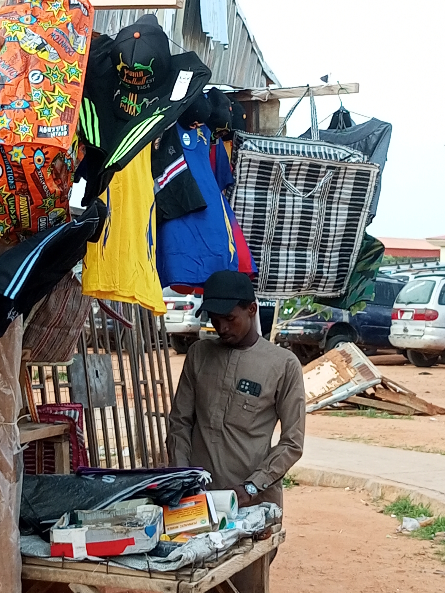 This media file captures an outdoor market scene in Gombe, Nigeria. The primary subject is a young man wearing a light brown, long-sleeved tunic and a black baseball cap. He stands behind a wooden stall laden with various goods. The stall displays an assortment of items including several black baseball caps with logos, bright orange fabric with a repeated pattern of eyes and stars, yellow and blue t-shirts, and a large checkered bag made of black and white fabric. Some items are hung on display, while others are stacked on the stall.
The man appears to be a vendor, possibly attending to his merchandise or interacting with a customer, though no other people are clearly visible in the immediate vicinity. He has a wristwatch on his left wrist and a dark-colored object, possibly a phone, tucked into the breast pocket of his tunic.
In the background, the setting includes several vehicles, including what appear to be SUVs or vans, parked on a dirt or gravel surface. There's also a hint of buildings with corrugated metal roofs and some wooden structures. The overall impression is that of a bustling, informal marketplace.
The time of day seems to be daytime, indicated by the bright natural light illuminating the scene. The weather appears to be fair, with no visible rain. The ground is dusty, characteristic of many outdoor markets in dry climates.
There is visible text on some of the items, including on the orange fabric where "DOWNTOWN" is repeated. Some of the caps also have logos and text, such as "PUMA" and "FOOTBALL EST 1948". On a yellow t-shirt, faint blue text is visible. On the stall, a box with red and white markings and some other packaged goods are present. A yellow box with text that includes "PERMANENT MARKER" is also visible. The scene evokes a sense of everyday commerce and activity.