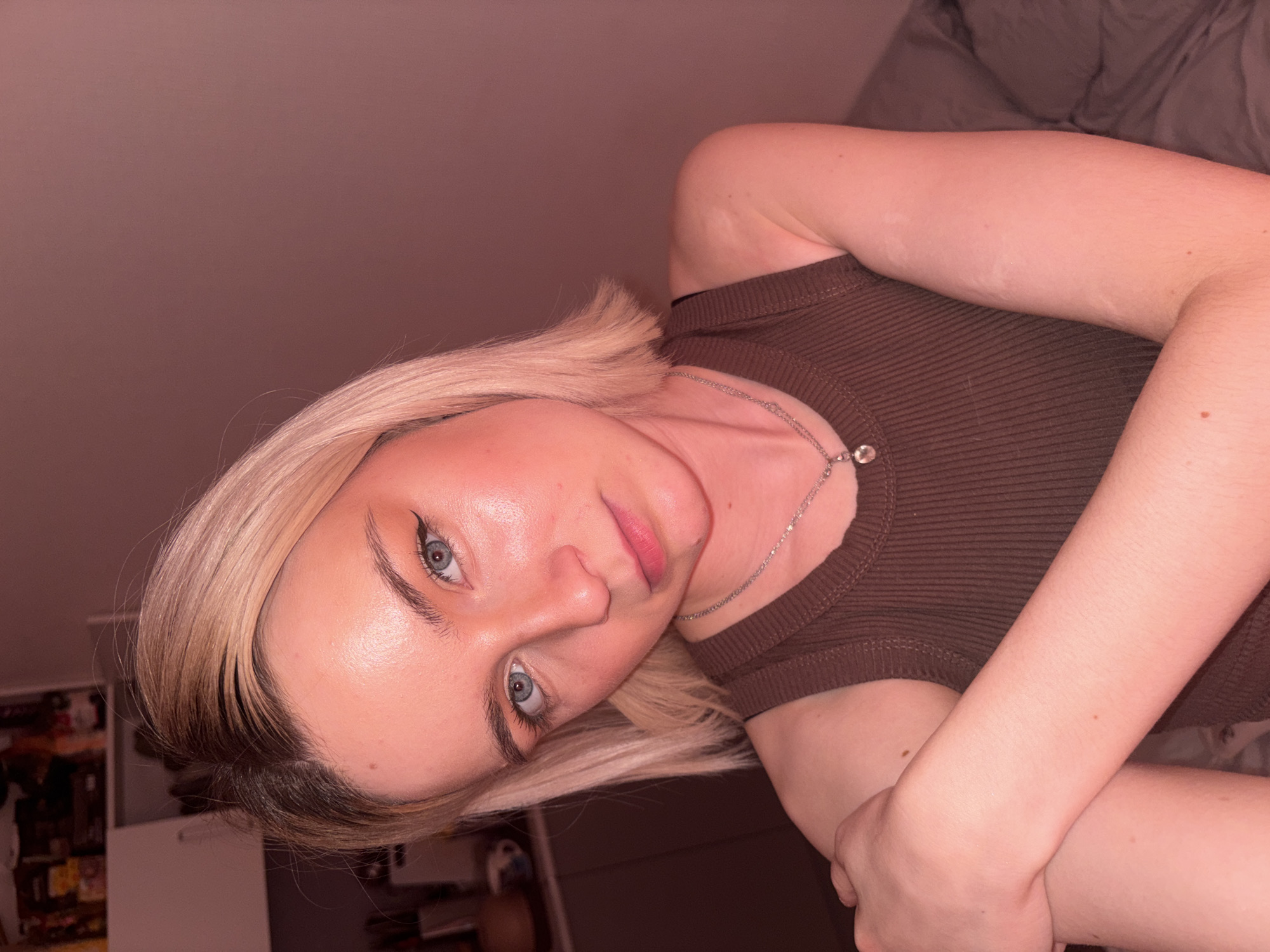 The image is a rotated, close-up portrait of a young woman, appearing to be a selfie. She has fair skin, striking light blue eyes, and blonde hair with visible dark roots, cut in a bob style that frames her face. Her head is slightly tilted, and she gazes directly at the camera with a neutral to subtly pensive expression, her lips gently parted in a slight pout. She wears noticeable winged eyeliner and has well-defined eyebrows. Her skin has a luminous or dewy finish.
She is dressed in a dark brown, ribbed tank top or camisole and wears a delicate silver chain necklace with a small, sparkling pendant. Her arms are crossed in front of her, resting close to her body.
The background suggests an indoor environment. To her upper left, a plain, muted reddish-brown wall is visible. To her lower right, a crumpled grey fabric, possibly a blanket or bedding, is out of focus. Further in the blurred background on the far left, there are faint outlines of shelves or furniture. The overall lighting is warm, indicating artificial light, possibly from an indoor evening setting. There is no visible text in the image.