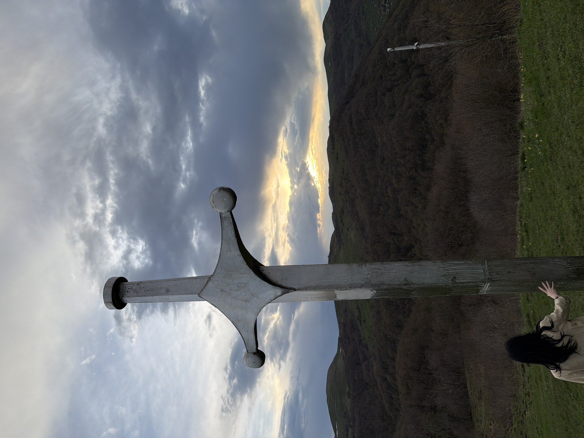The image shows a large metal sword monument in Zemo Nichbisi, Georgia. The sword is in the foreground, pointing diagonally toward the sky. A mountain range is in the background with sunlight breaking through the clouds. A person with long dark hair and a tan jacket is reaching out toward the base of the sword on the right side of the image. Green grass is visible near the person and the base of the sword. The sky is filled with grey and white clouds, with a section where sunlight is shining through. A wooden cross-like structure is on a hill in the background.