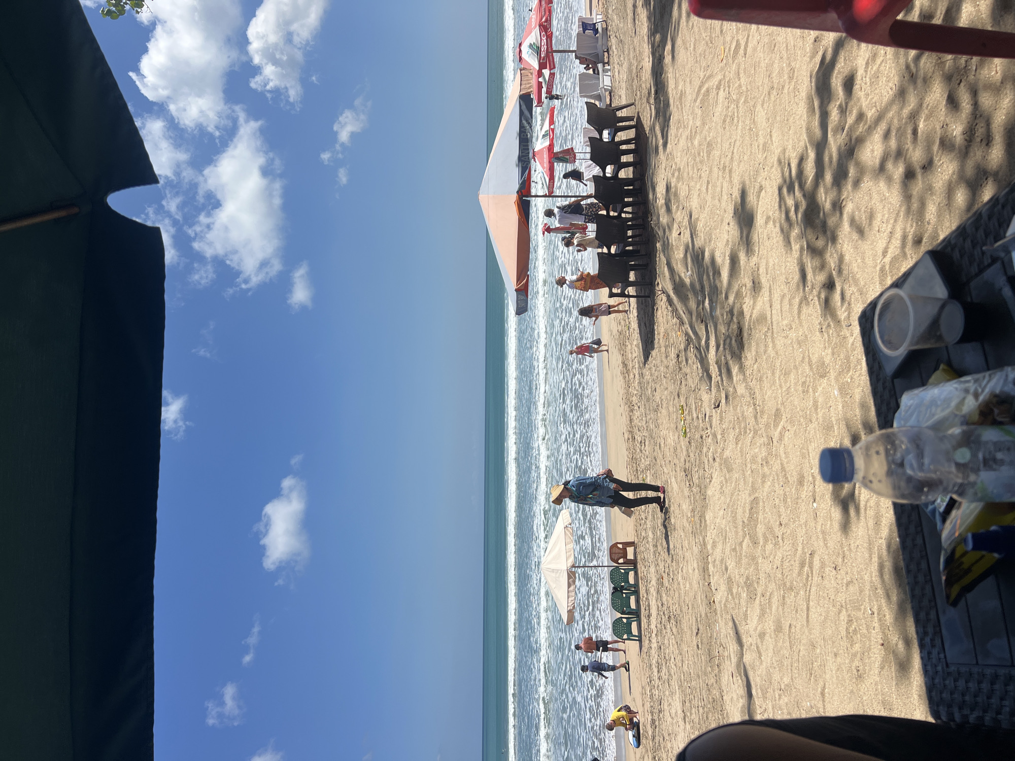The image depicts a sunny day at Kuta Beach, Bali, Indonesia. The foreground shows a table with a water bottle and a plastic cup, suggesting a relaxing break from the beach. The middle ground is the sandy beach itself, with several beach umbrellas and chairs set up for rent or use. People are scattered across the beach; some are near the water, while others are closer to the umbrellas. A woman in a hat and denim shirt is standing on the sand, and further down the beach, there are children and adults engaging in various activities. The ocean stretches out to the horizon, with gentle waves rolling onto the shore. The sky is a clear blue with a few fluffy white clouds. The overall atmosphere is one of leisure and a typical beach day in Kuta.