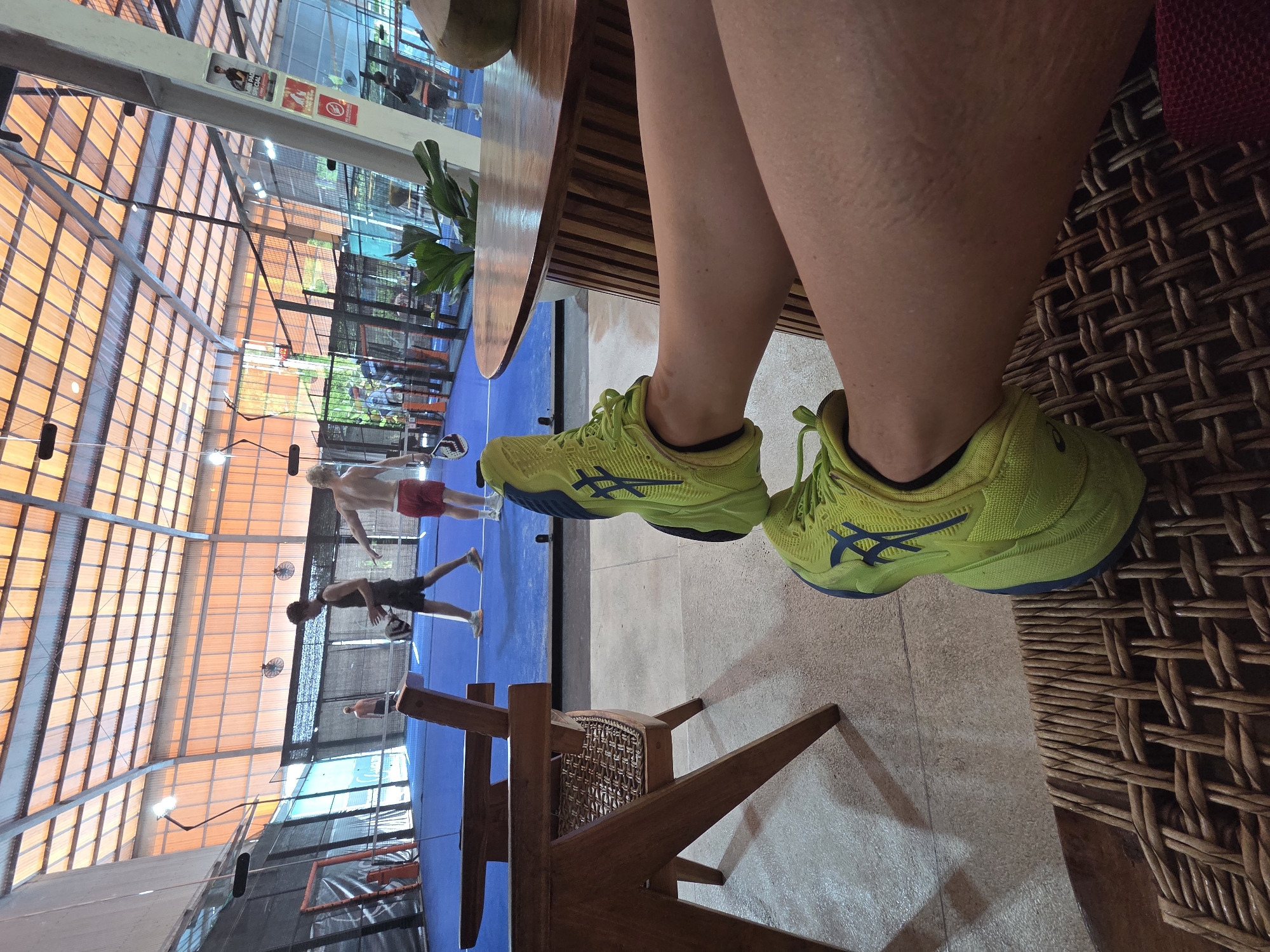 The photograph depicts a pair of legs wearing lime green athletic shoes with blue accents, resting on a woven rattan chair. The legs are fair-skinned. The setting appears to be a recreational facility, specifically a padel tennis club, as evidenced by the padel courts visible in the background, where three men are engaged in a game, wearing minimal clothing. The court is illuminated by natural light filtering through a glass ceiling with a honeycomb-like pattern. The scene also includes a wooden table adjacent to the chair. The location is Unknown, Unknown.