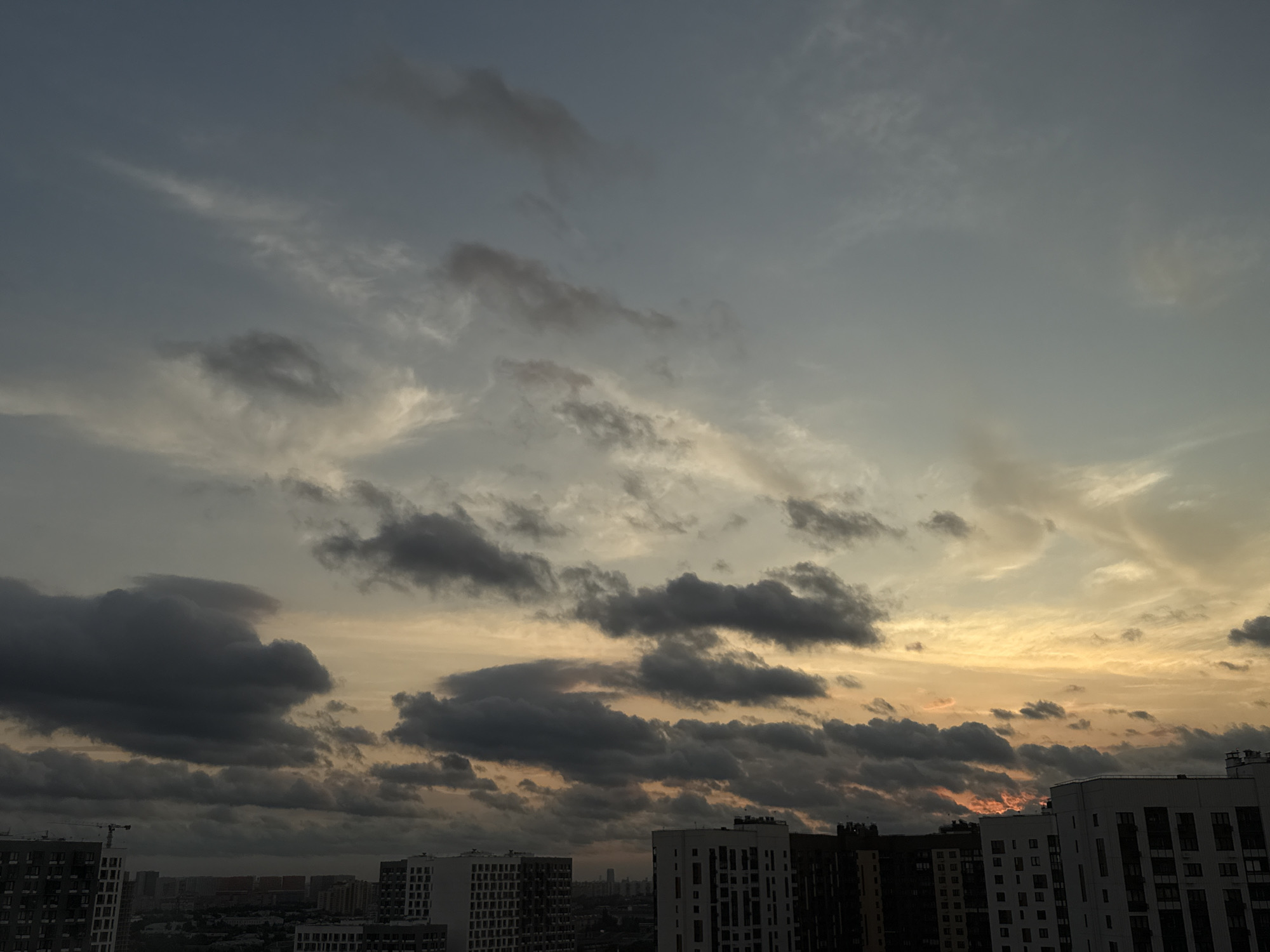This is a daytime, outdoor, medium shot photograph taken from a high vantage point, likely a balcony or rooftop, looking out over a city. The sky dominates the upper two-thirds of the frame, filled with a dramatic sunset. Dark, heavy clouds are scattered across the sky, with lighter, wispy clouds also visible. The setting sun casts a warm, orange glow through gaps in the clouds, illuminating the undersides of some of the cloud formations.
The lower third of the image shows a cityscape. Several large, modern apartment buildings are visible, characterized by their multi-story structures and geometric facades. Some buildings are lighter in color, appearing white or light gray, while others are darker. In the far distance, more buildings and a hazy cityscape are visible, suggesting a densely populated urban area. A construction crane can be seen near the left side of the image, indicating ongoing development. The overall atmosphere is one of twilight in an urban setting. The location is specified as Lyubertsy, Russia.