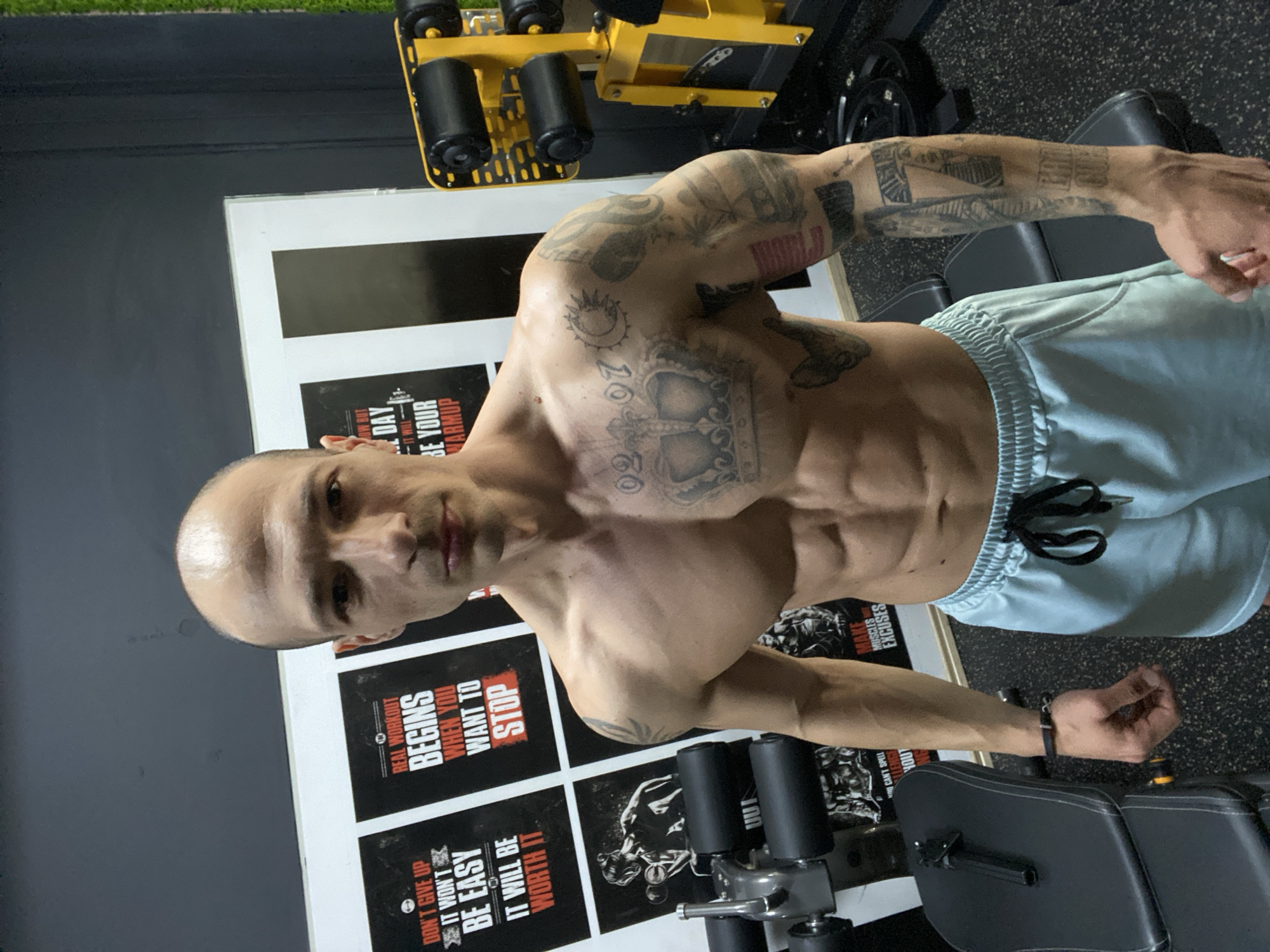 The image, taken in Pleven, Bulgaria, displays a muscular man posing in what appears to be a gym or fitness studio. The man, who is shirtless and has a shaved head, exhibits a lean and well-defined physique with visible abdominal muscles. He is extensively tattooed with black ink designs covering his chest, shoulders, and arms, including a prominent crown on his chest and a sun symbol on his right shoulder. He wears light blue athletic shorts with a black drawstring and has a neutral to serious expression while looking towards the camera.
The background features a dark grey wall adorned with several white-framed motivational posters. Visible gym equipment includes a yellow and black machine with padded rollers in the upper part of the frame, and a black padded bench or seat in the lower right. The floor appears to be dark, textured gym flooring. The lighting suggests an indoor setting.
Visible text on the motivational posters includes:
* "DON'T GIVE UP"
* "IT WON'T BE EASY"
* "IT WILL BE WORTH IT"
* "REAL WORKOUT BEGINS WHEN YOU WANT TO STOP"
* "TODAY, IT WILL BE YOUR WARMUP"
* "MAKE MUSCLES EXCUSES"