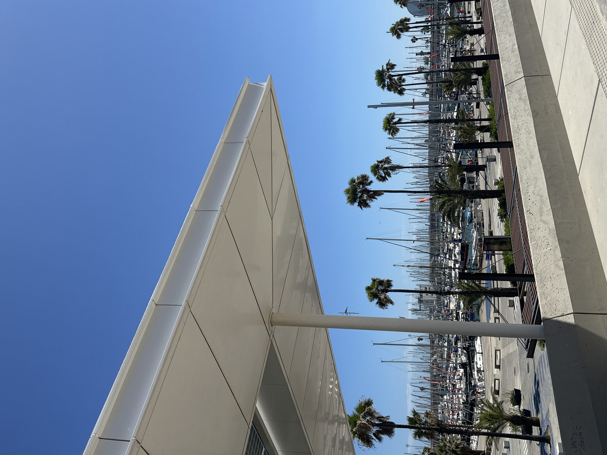 The image shows a modern, angular building in Barcelona, Spain, under a clear blue sky. A row of palm trees lines a promenade or walkway, with a marina filled with sailboats visible in the background. The building is predominantly white with clean lines and appears to be constructed of modular panels. A cylindrical column supports a section of the building. The palm trees are aligned neatly along the walkway, which is made of grey concrete. The sailboats in the marina have masts that stretch towards the sky. The photograph is taken from a low angle, emphasizing the height of the building and palm trees.