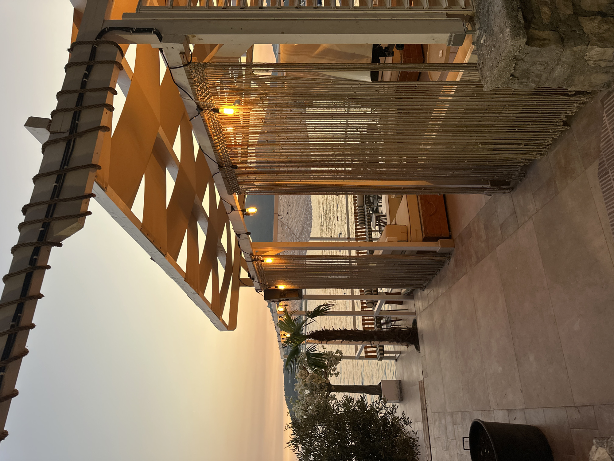 The image depicts an outdoor seating area of a beach bar or restaurant, likely located in Budva, Montenegro. The scene is set at dusk, with the sky displaying warm orange and yellow hues characteristic of a sunset. The foreground is dominated by a tiled patio leading towards a covered structure. This structure features a slatted roof with fabric awnings creating a patterned effect. Decorative ropes are strung along the top beams. String lights with Edison-style bulbs are hung across the structure, casting a warm glow. A macrame-like curtain made of thick rope hangs as a partition.
Beyond the structure, the calm sea is visible, with a distant coastline and hills. Several palm trees, including a prominent one in the middle ground, are present, adding to the tropical or Mediterranean ambiance. Seating arrangements, including tables and chairs with striped cushions, are visible on a raised platform near the water's edge. An olive tree with white decorative blossoms is situated on the left side of the image. A dark-colored trash bin is placed on the patio. The overall atmosphere is serene and inviting, suggesting a peaceful evening by the sea. There are no people visible in the image.