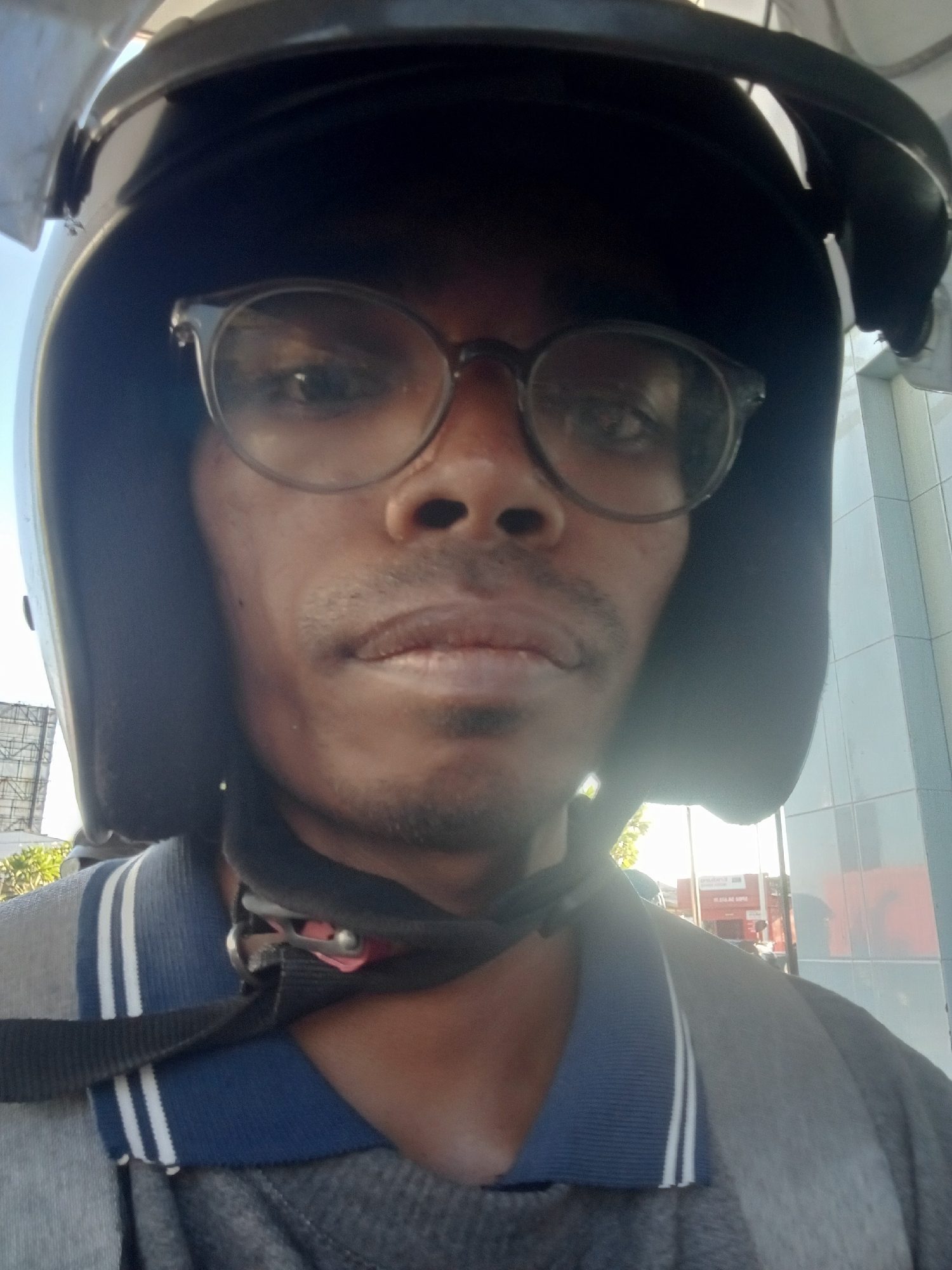 A close-up, forward-facing selfie of a Black man wearing a dark helmet and clear-framed glasses. He has a light mustache and goatee. He is wearing a collared shirt with blue and white stripes on the collar. The background is out of focus, but appears to be an urban environment with a tiled building on the right and some trees and a billboard structure on the left. The overall lighting suggests it is daytime. The man is likely in Sawotratap, Indonesia, based on the metadata.