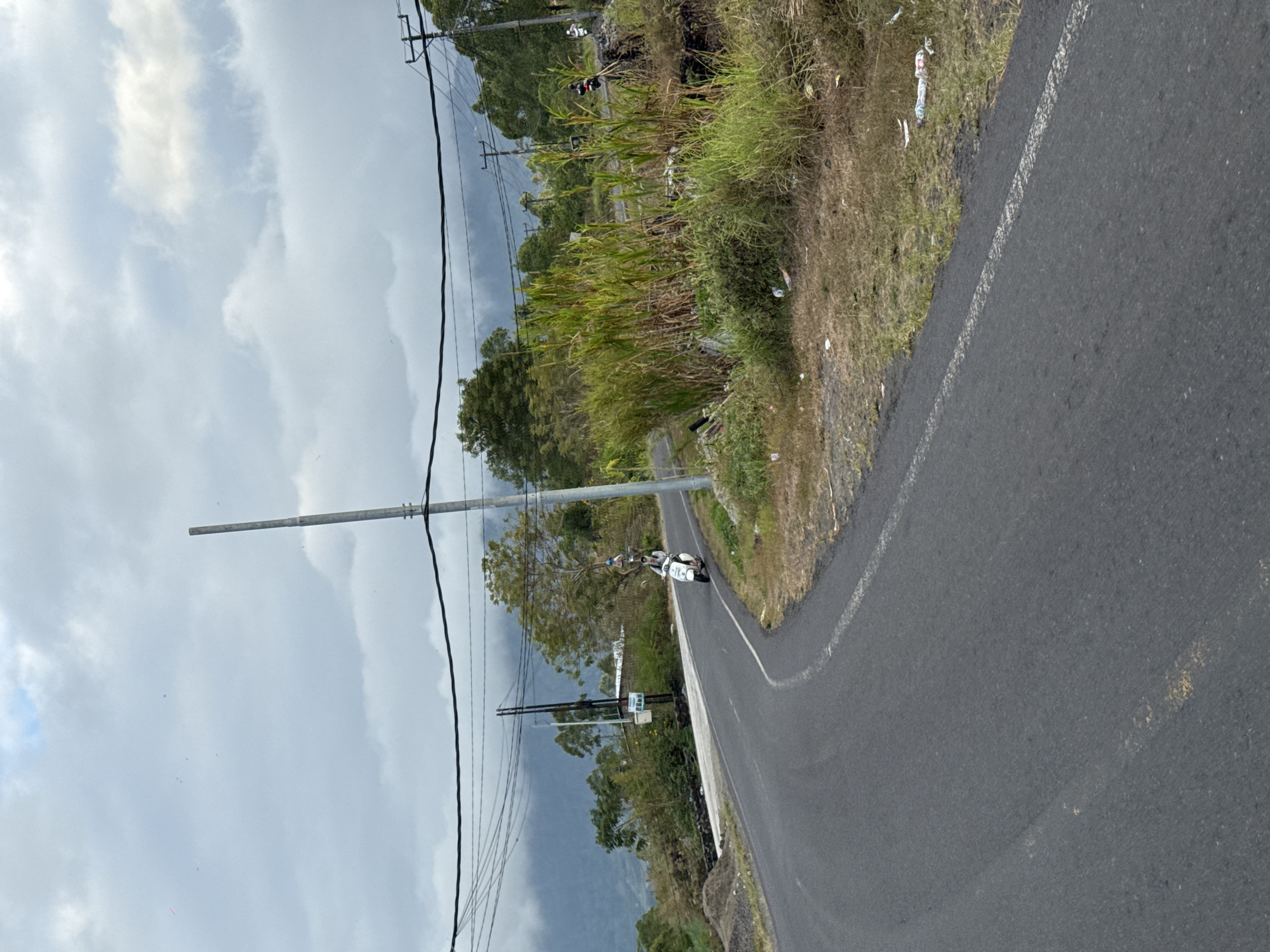 The image depicts a scenic road winding through the mountainous landscape of Kintamani, Indonesia. The road is paved and curves to the left, with a white line marking its edge. On the right side of the road, tall grasses and vegetation grow, interspersed with some scattered litter, including plastic bottles. A tall, weathered utility pole stands prominently on the right side of the road, with power lines stretching across the frame. In the background, lush green trees and hills rise towards a cloudy sky. Further in the distance, mountains are visible, shrouded in mist, suggesting a cool and humid climate. A white scooter with a rider is captured in motion on the road, heading away from the viewer. Another scooter is visible further down the road, also moving away. The overall impression is of a rural, somewhat remote location with ongoing transportation activity.