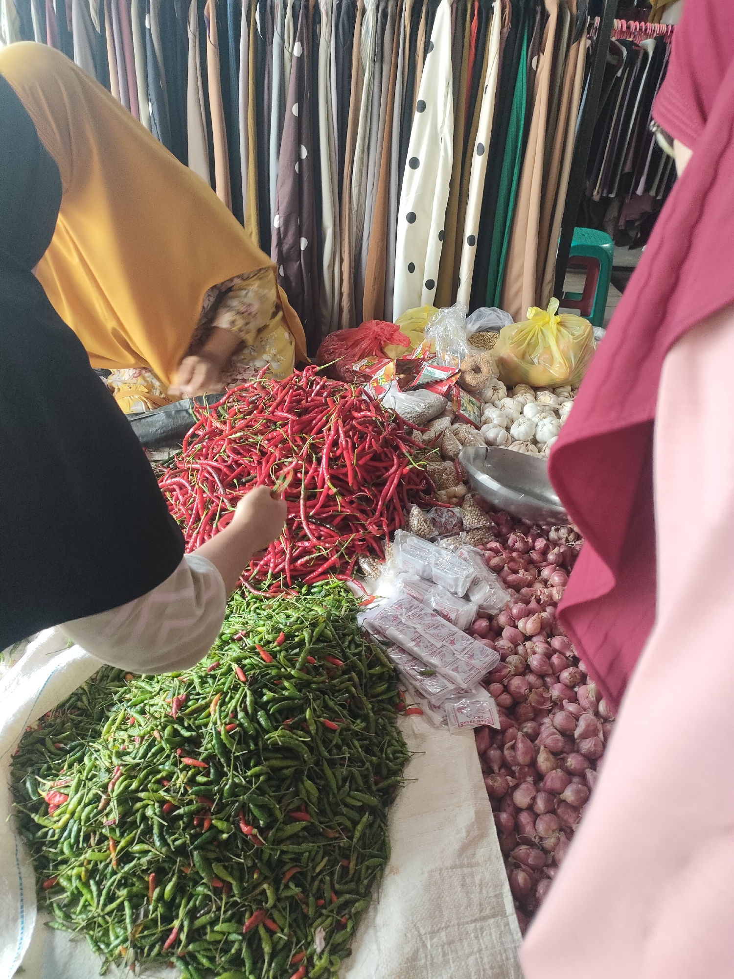 A market stall in Sukoyoso, Indonesia, displays a variety of produce and goods. In the foreground, a pile of small, green chilies is visible in a white sack. To the right of the chilies, a large quantity of red onions is arranged on a light-colored surface. Behind the onions, a metal bowl sits atop a heap of garlic bulbs. Further back, a large pile of red chili peppers is present. Several plastic-wrapped packages, likely containing spices or seasonings, are scattered amongst the produce. In the background, racks of colorful fabrics are displayed. Two individuals are partially visible. One person, wearing a yellow headscarf and patterned clothing, is reaching towards the red chilies. Another person, wearing a dark hijab and light-colored sleeves, is also near the chilies, with their hand reaching out. A third person, wearing a maroon hijab and pink garment, is on the far right, their back to the camera. A small green plastic stool is visible behind the produce. The scene suggests a bustling market environment.