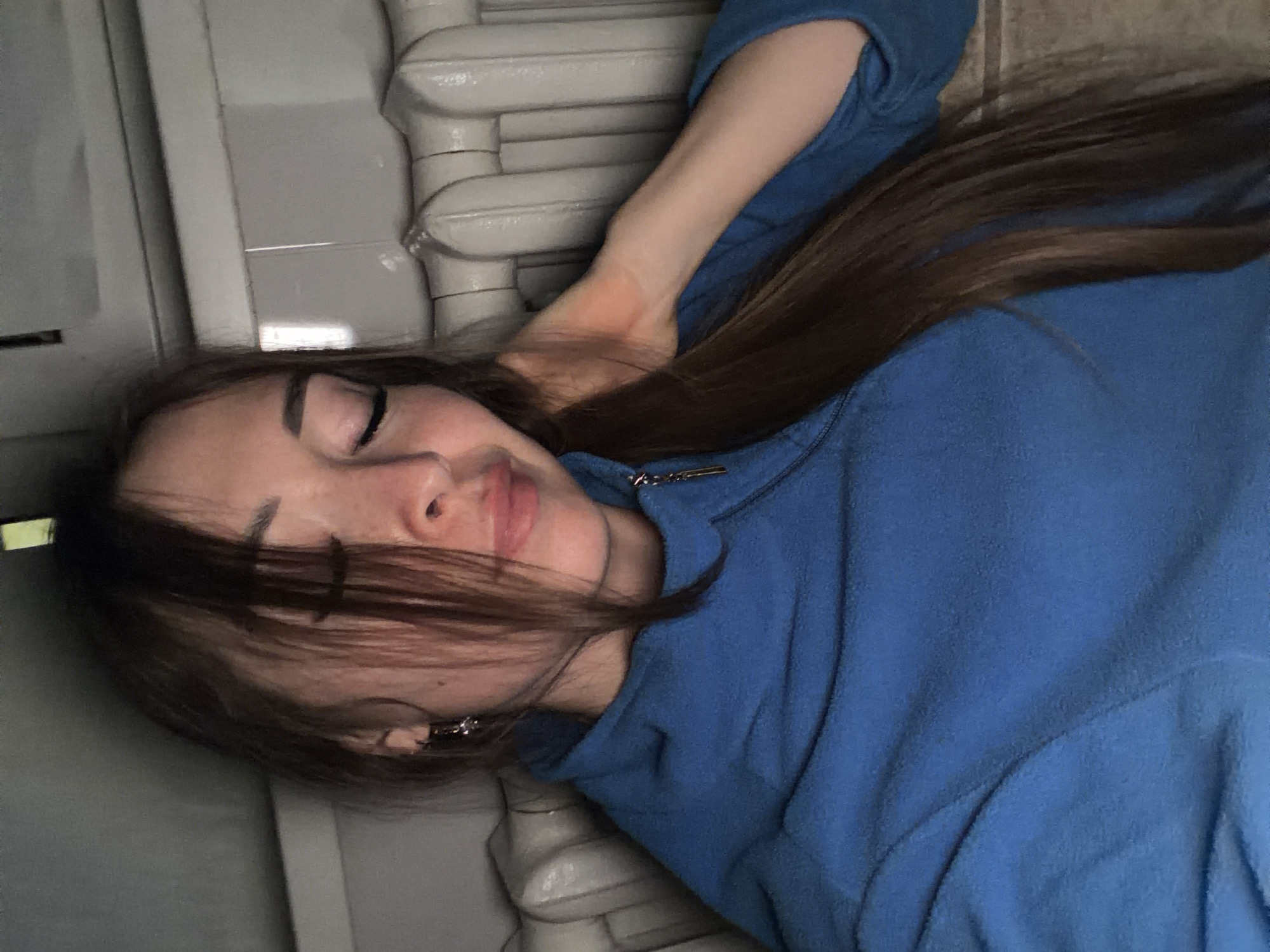 The image shows a young woman lying down, possibly asleep or resting. She is wearing a blue hoodie. Her eyes are closed, and her head is tilted. Her long brown hair is spread around her face and shoulders. She has freckles on her face and is wearing small earrings. The background appears to be indoors, with a light-colored radiator and a wall that seems to be painted or tiled in neutral tones. The overall lighting suggests it could be daytime, but the interior setting makes it difficult to determine the exact time or weather conditions. The mood conveyed is one of relaxation and quiet. There is no visible text in the image. The location context provided is Япрыково, Russia.
