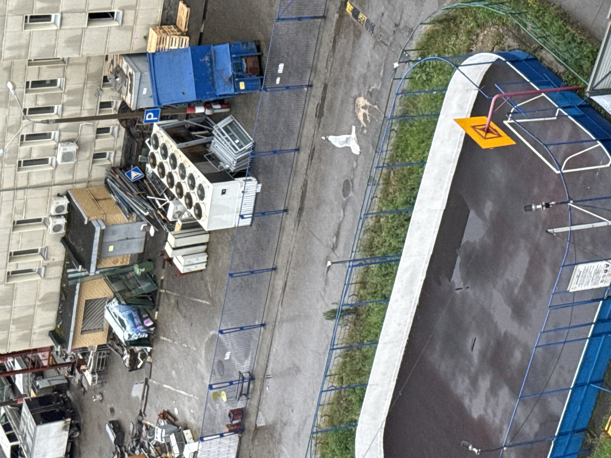 The image shows an outdoor scene, likely in an industrial or storage area, with a basketball court in the foreground. The basketball court is partially wet and has a single basketball hoop with an orange backboard. A dark-colored fence encloses the court. Beyond the court, there is a paved area with various objects, including construction materials, large white appliances, a blue skip, and a stack of wooden pallets. A white figure, possibly a person in protective clothing or a large piece of white plastic, is lying on the ground in this area. In the background, a multi-story building with numerous windows and air conditioning units is visible. Some areas of the paved ground appear wet. This scene is located in the city of Nizhny Novgorod, Russia.