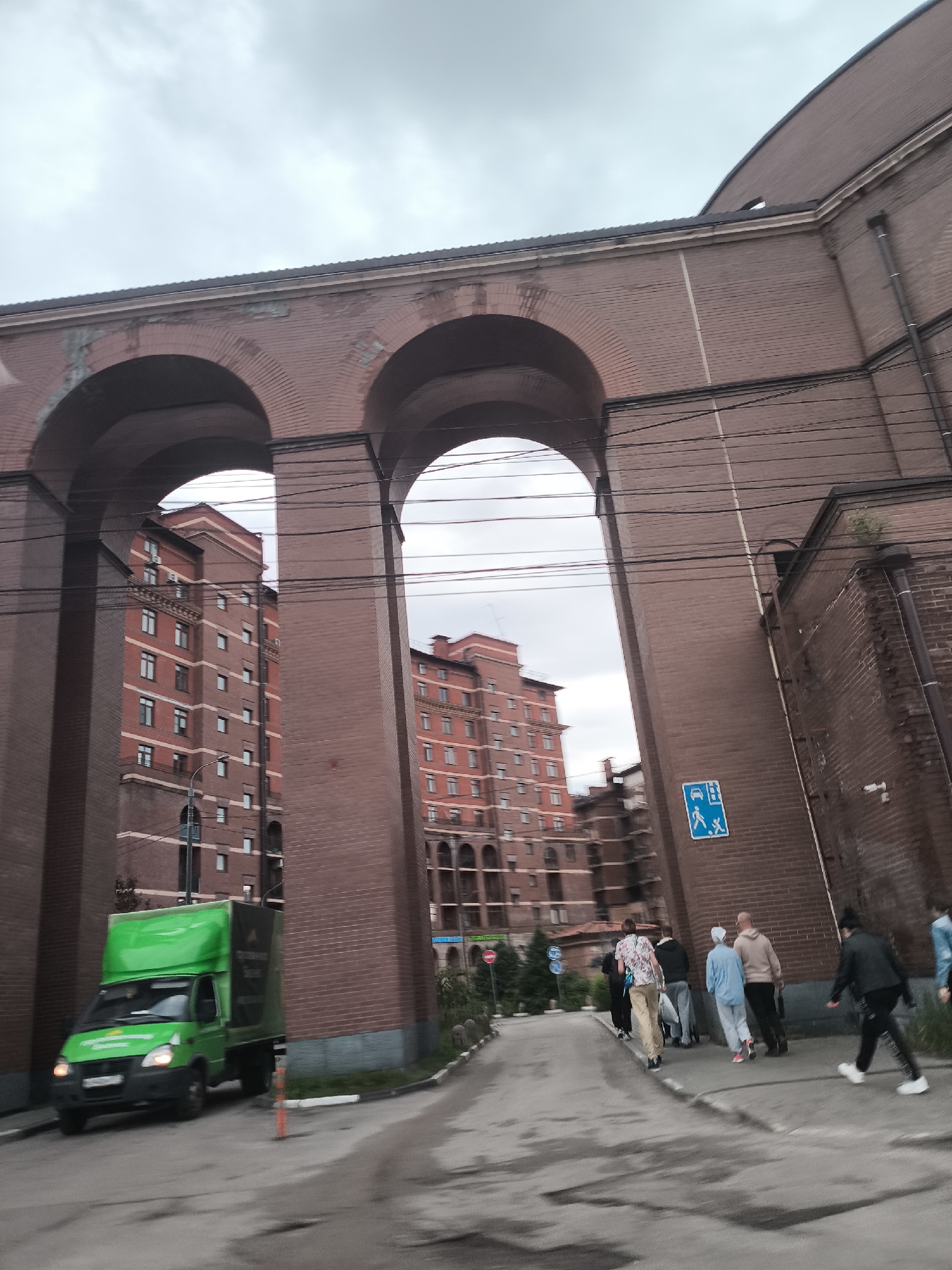 The image captures a street scene under a cloudy sky, featuring a large brick structure with two prominent arches. In the foreground, a green delivery truck is parked on the left. To the right of the truck, a group of people are walking away from the camera on a paved pathway. Behind the arches, a tall, red brick apartment building is visible. A blue and white traffic sign is mounted on the brick structure, indicating a pedestrian crossing. The overall impression is an urban environment with architectural elements and everyday activity.