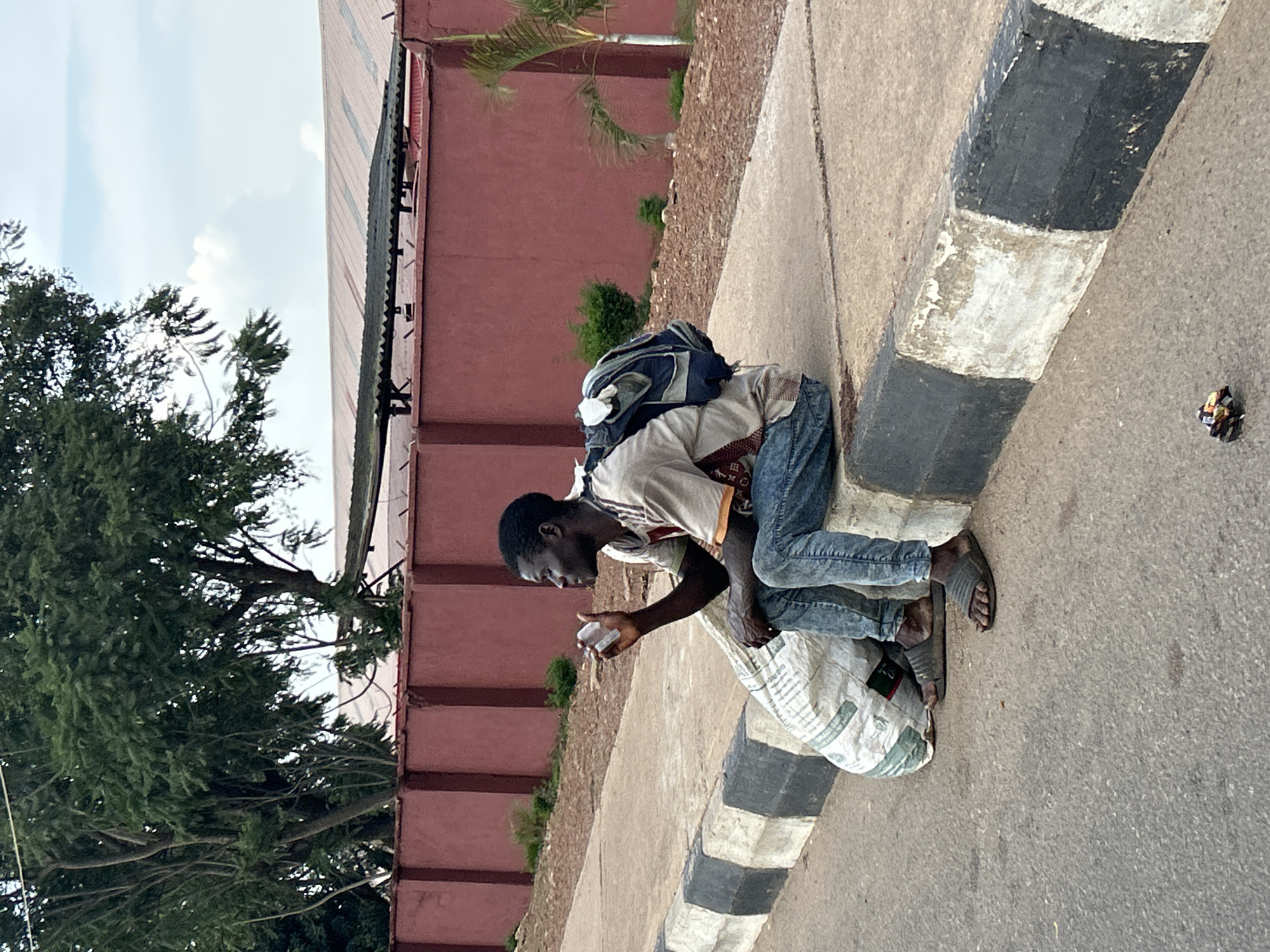 A man sits on a concrete curb in Jos, Nigeria, holding a small object, possibly a phone, to his ear. He is dressed in a light-colored t-shirt, blue jeans, and dark sandals. A blue and gray backpack is on his back, and a large, white, woven sack rests beside him on the curb. The curb is painted with black and white stripes. Behind the curb, a red stucco wall runs horizontally across the frame, topped with a dark corrugated roof. Trees with green foliage are visible behind the wall, and the sky above is partly cloudy. A palm tree stands to the right of the frame. The ground in front of the curb is paved asphalt.