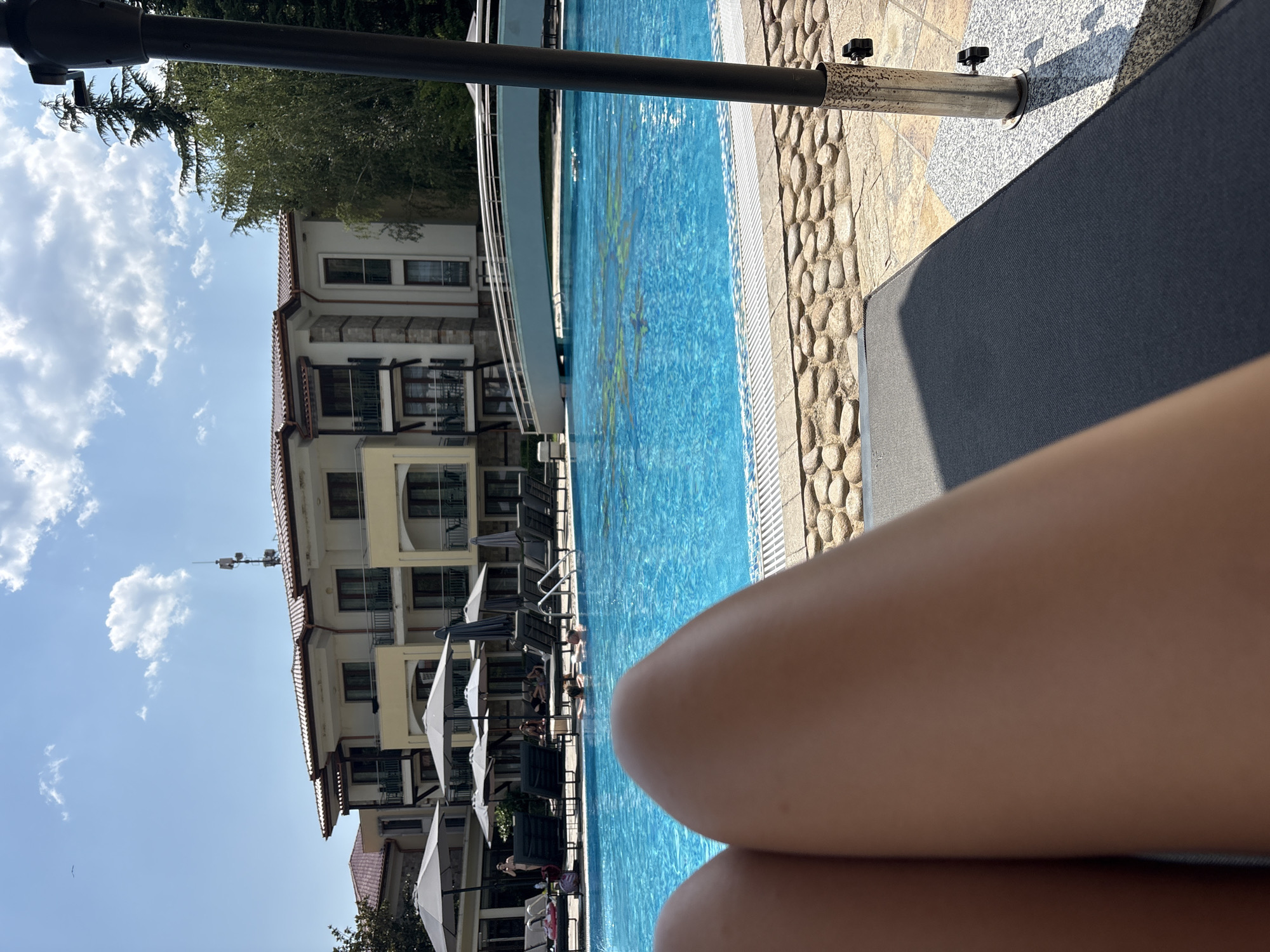 A first-person perspective shot shows a person's legs resting on a dark grey lounge chair. In the background, a bright blue swimming pool is visible, with people swimming and relaxing. Several lounge chairs and umbrellas are arranged around the pool. Beyond the pool, a light-colored building with multiple stories and balconies is visible. A white pedestrian bridge arches over a section of the pool. The sky is blue with scattered white clouds, and trees are present on the right side of the frame. The ground around the pool is paved with small stones.