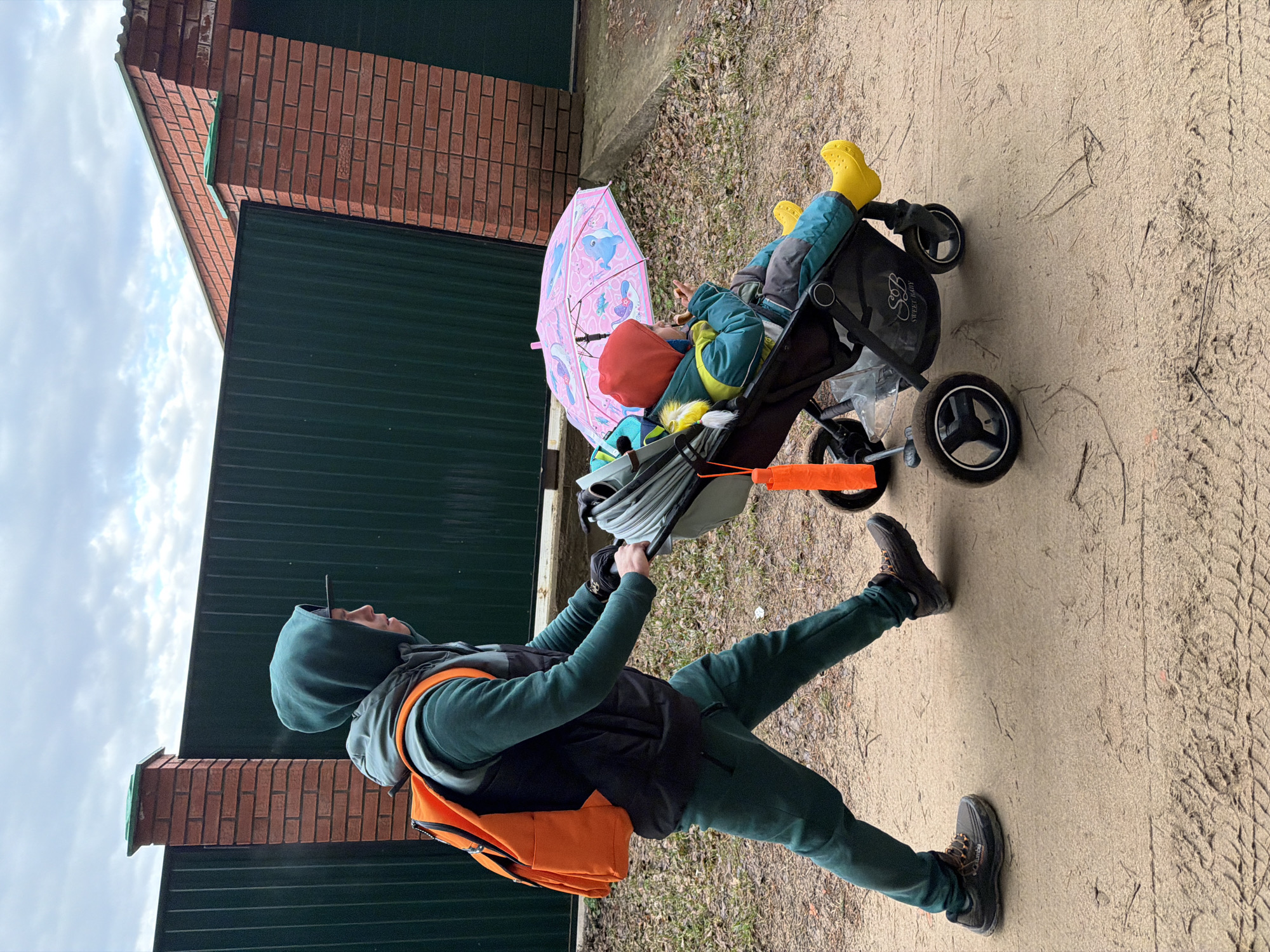 An outdoor photograph, rotated to be upright, captures an adult pushing a child in a stroller along a sandy path in Chaikovsky, Russia. On the left, an adult, likely male, is dressed in a dark green hooded tracksuit, a black vest, and an orange backpack. He wears dark boots, one of which bears the "Columbia" brand logo. He is actively pushing a dark-colored stroller.
Seated in the stroller on the right is a young child, wearing a blue jacket, a red hat, and distinctive bright yellow rain boots. The child appears calm, possibly eating a small snack, with a pink umbrella featuring cartoon dolphins and other sea creatures positioned beside them. The stroller has "SWEET BABY" written on its frame and is equipped with robust wheels, navigating the unpaved, sandy ground strewn with dry leaves and debris.
In the background, behind the subjects, is a building or enclosure. Its lower section consists of a dark green corrugated metal fence or wall, topped by a red brick structure. The sky above is overcast with white and grey clouds, suggesting a cloudy day. The presence of the umbrella and the child's rain boots hints at recent rain or the potential for it. The scene depicts a casual outdoor stroll.