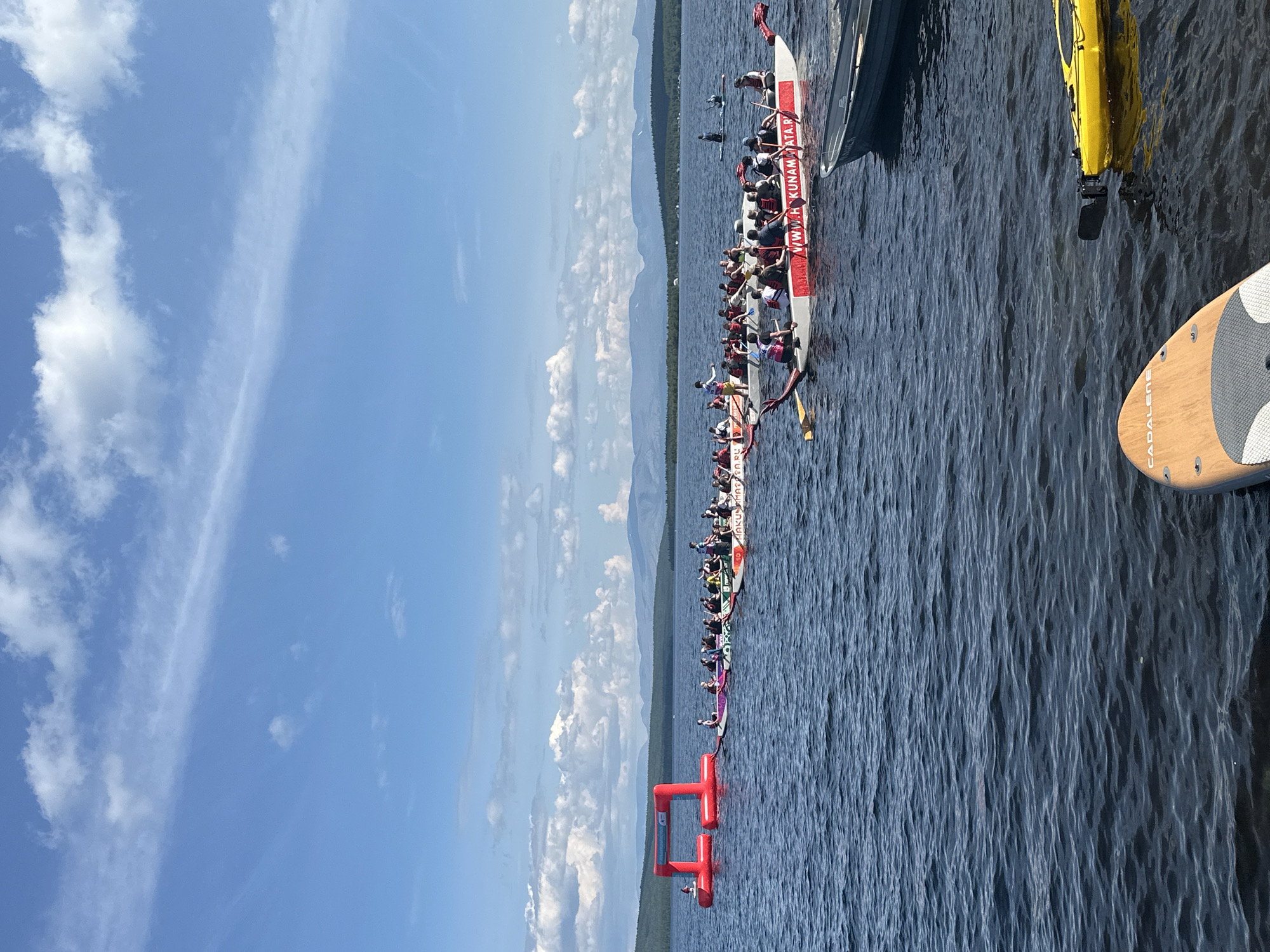 The image depicts a sunny day on a large body of water, possibly a lake, in Monchegorsk, Russia. In the foreground, a paddleboard is partially visible, along with a yellow kayak. The middle ground features several dragon boats, each filled with numerous participants wearing life vests and holding paddles. One of the dragon boats has red lettering on its side that appears to be a website address, "WWW.H KUNAMATA.P". To the left, a red inflatable archway structure is positioned on the water, suggesting a starting or finishing point for a race or event. In the background, a tree-covered shoreline is visible, with mountains and a blue sky with scattered white clouds forming the horizon. The overall scene indicates a water-based recreational activity or competition.