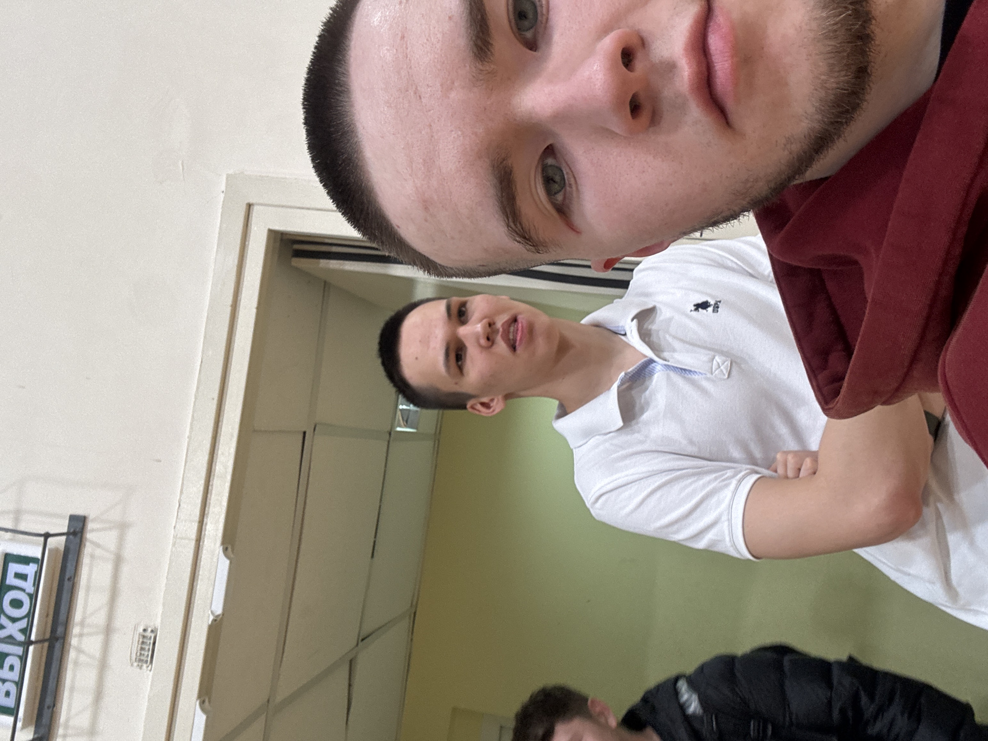 The image shows a selfie of two men in a hallway. One man is closer to the camera, partially obscuring the view of the other. Both men have short hair. The second man is wearing a white polo shirt. The location appears to be indoors in Kazan, Russia, based on the Russian word "ВЫХОД" (EXIT) visible in the background on a sign mounted on the wall near the door. A third person is partly visible at the bottom of the image. The environment seems to be a public space like a gymnasium or a school corridor.