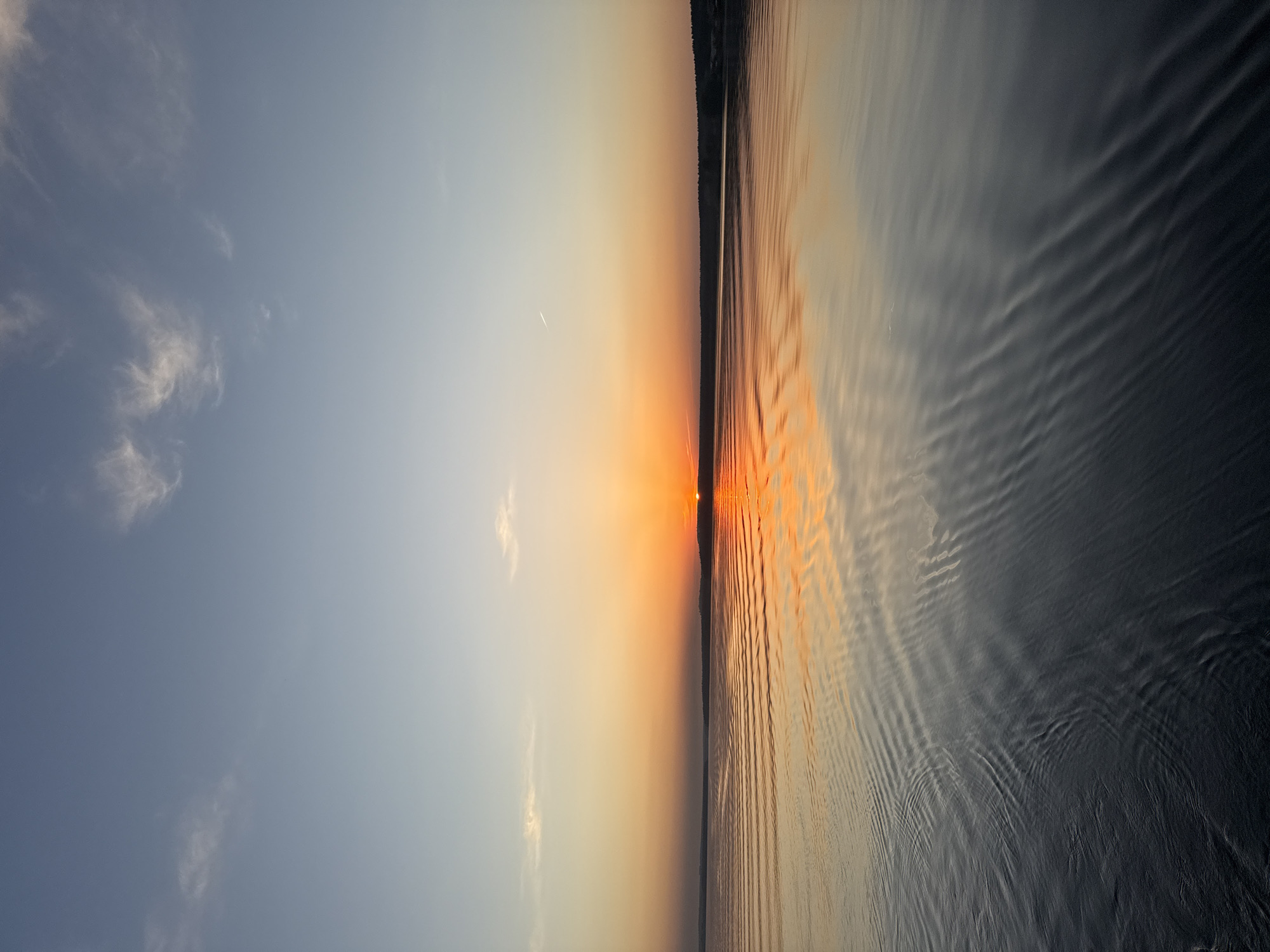 The image depicts a sunset over a body of water, likely a lake or reservoir. The sky transitions from a light blue with wispy clouds to a warm orange and yellow near the horizon. The sun is setting just above the horizon line, casting a bright orange reflection on the water. The water is calm with slight ripples, reflecting the colors of the sky. A dark strip of land, possibly a shoreline with trees, is visible in the distance along the horizon. Given the prompt, this image is likely taken in Votkinsk, Russia, and the body of water could be the Votkinsk Reservoir.