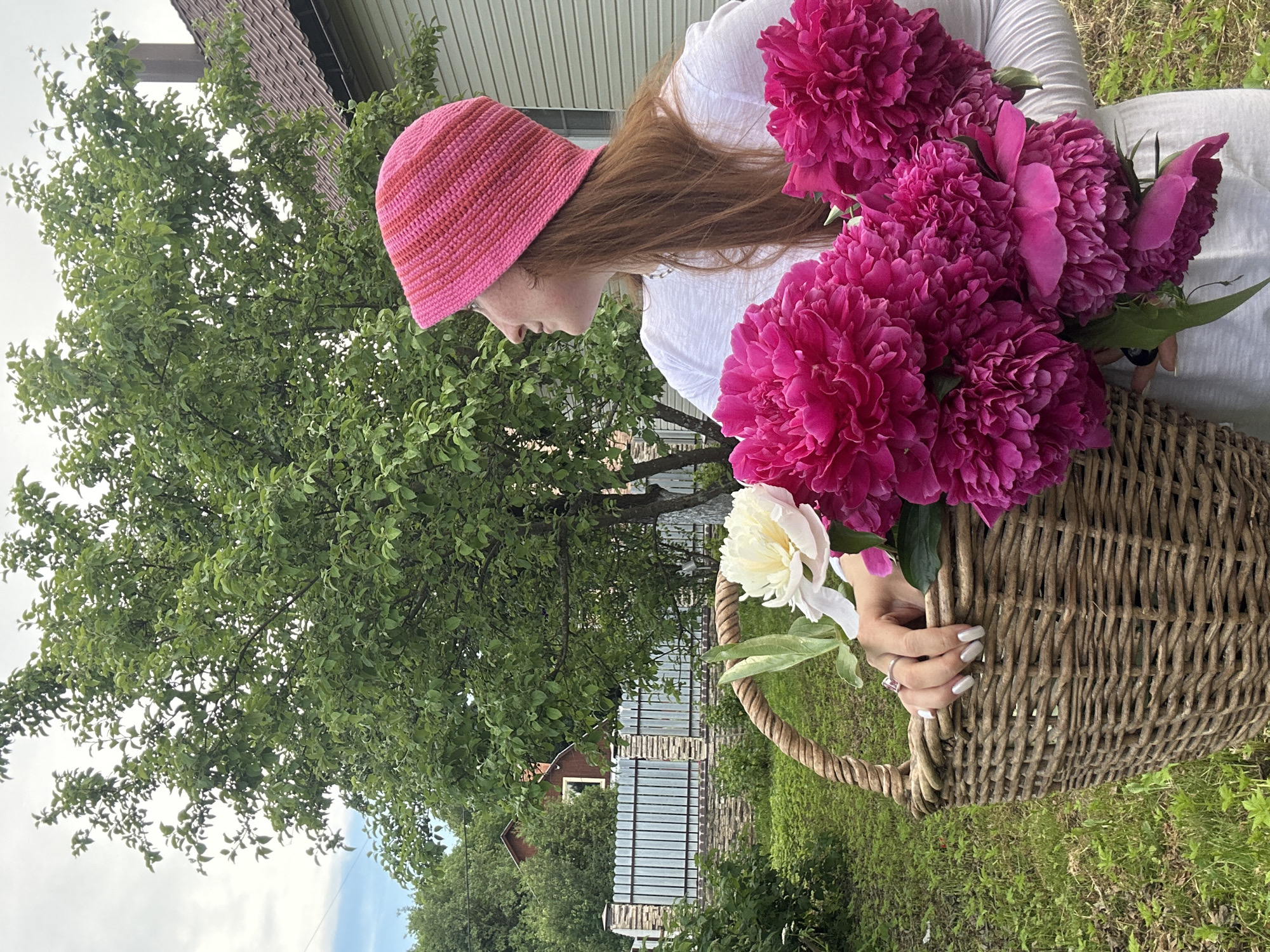 A woman with red hair and a pink knitted hat stands in a yard, holding a basket of pink and white peonies. She is wearing a white t-shirt and looking to the right. In the background, there is a house with a brown roof and a fence. The scene is set in a garden with green grass and trees. The location of Nizhny Novgorod, Russia is not discernible from the image.
