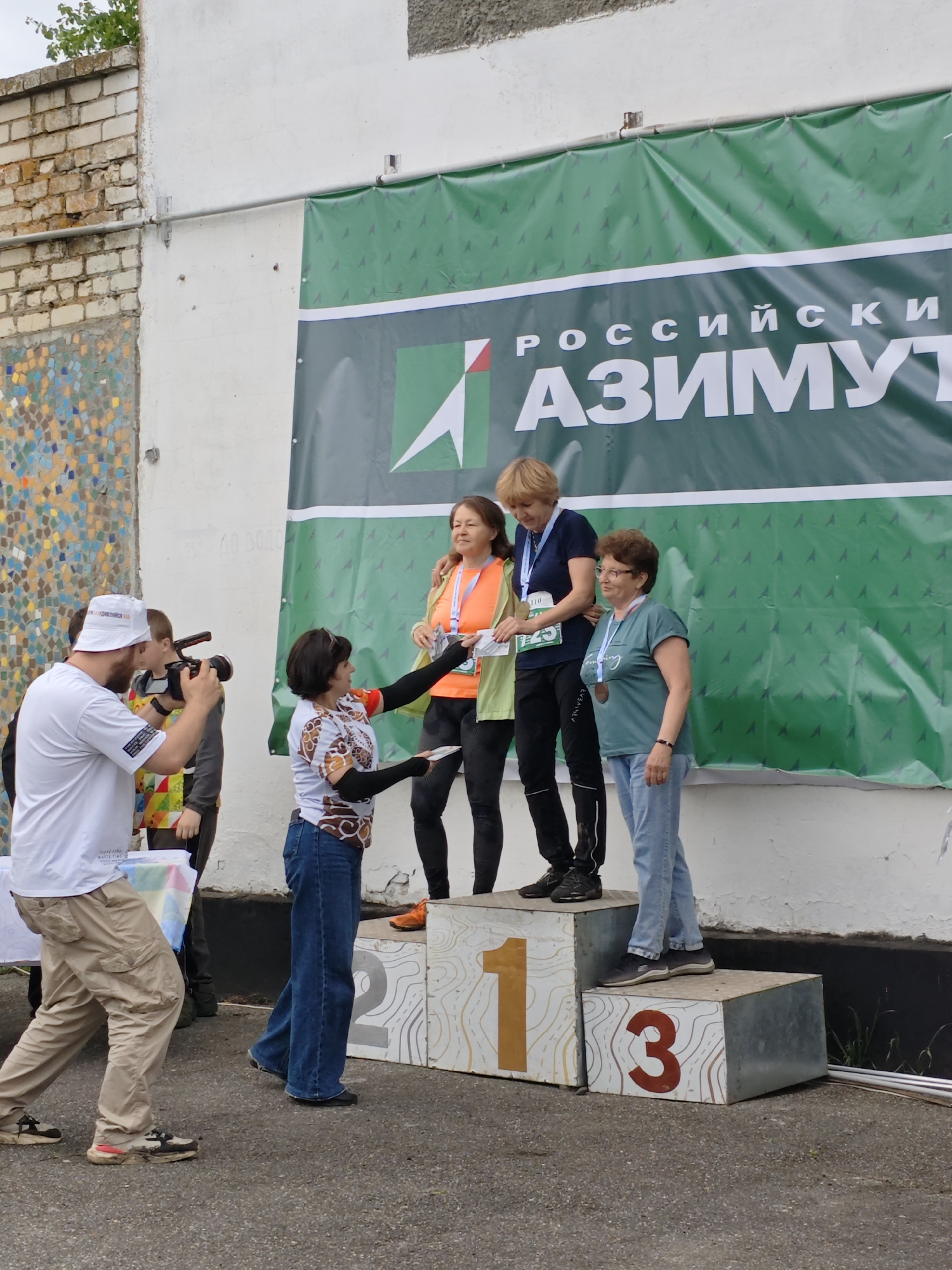 The image shows three women standing on a podium with the numbers 1, 2, and 3, indicating their ranking in an event. A woman standing in front of the podium is handing them papers. A man with a camera is taking pictures. Behind the podium is a green banner with the text "РОССИЙСКИЙ АЗИМУТ". The scene appears to be outdoors in Новоекатериновская, Russia. There is a white wall with some brick and colorful tilework visible behind the photographer.