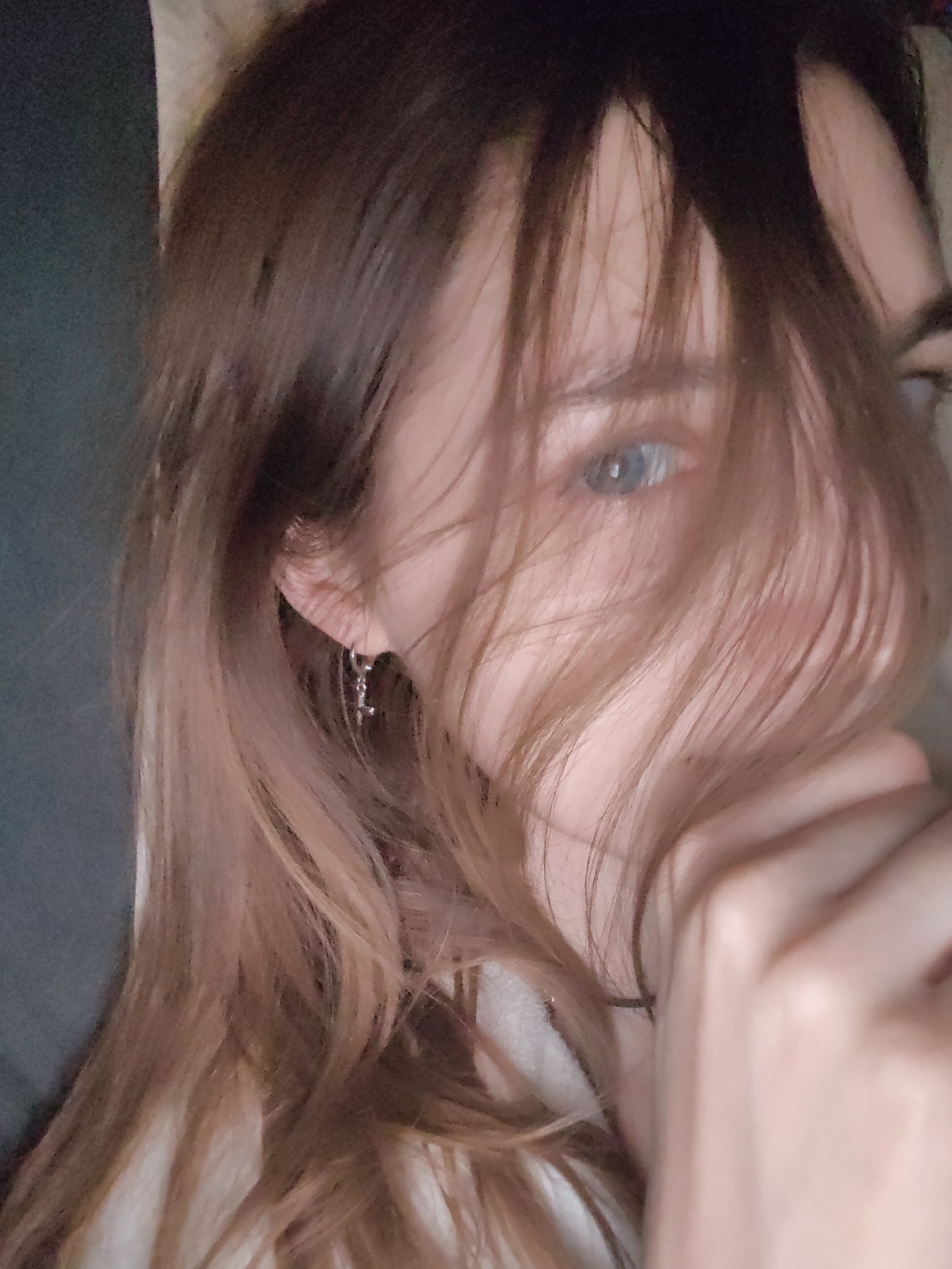 A portrait of a person with long, light brown hair partially obscuring their face. One eye is visible, appearing light blue. The person's hand is raised near their face, with fingers slightly curved. They are wearing an earring. The image appears to be taken indoors with muted lighting. There is no indication of location being in Chaikovsky, Russia.