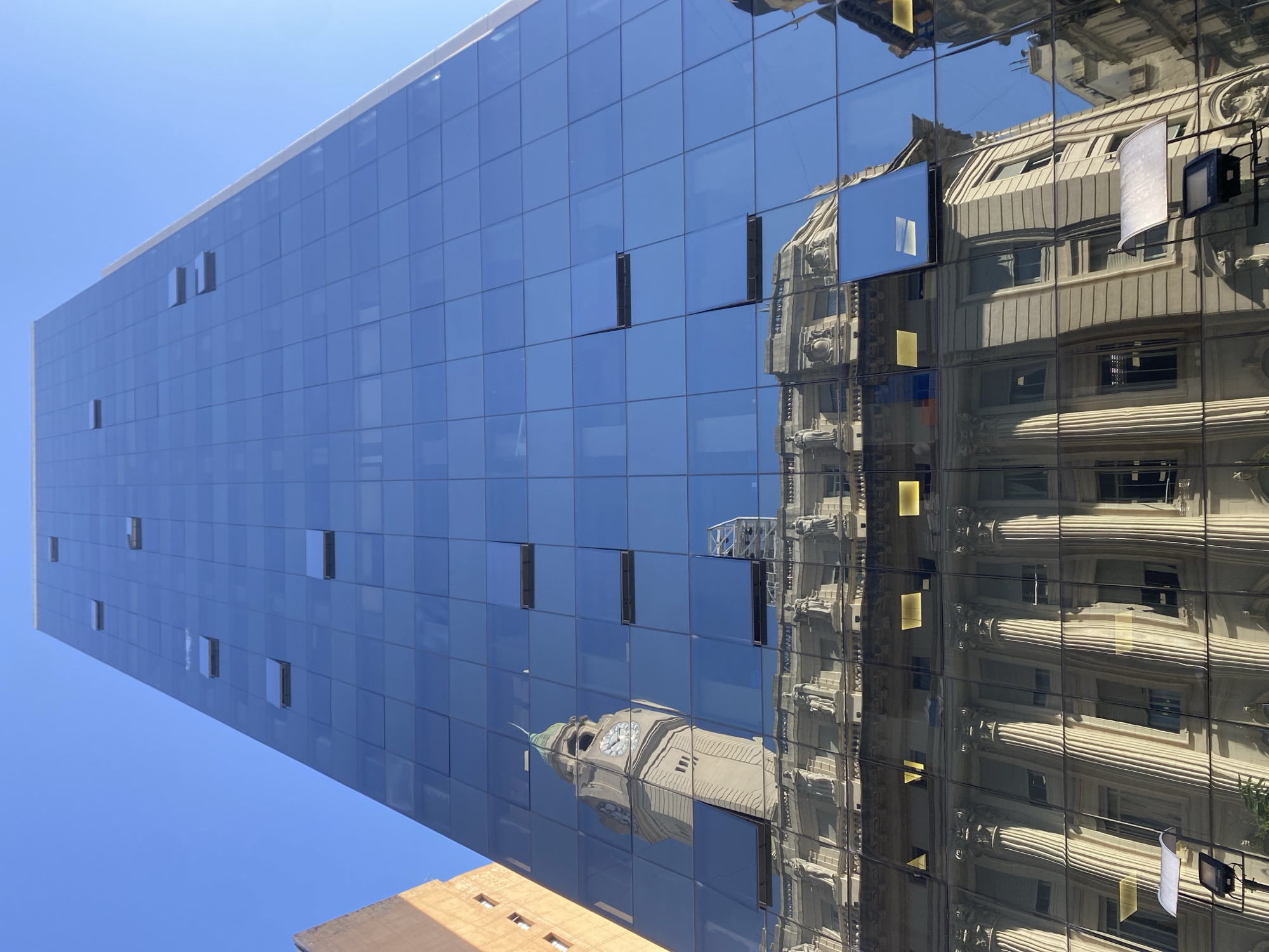 A low-angle shot captures the facade of a modern glass building. Its blue mirrored panels reflect the image of a classical-style building with architectural details like columns and ornate window frames, including a clock tower with a green roof. Some windows on the glass building are ajar. The sky is blue. This image is likely located in Buenos Aires, Argentina, due to the presence of classical buildings that are typical in the area.