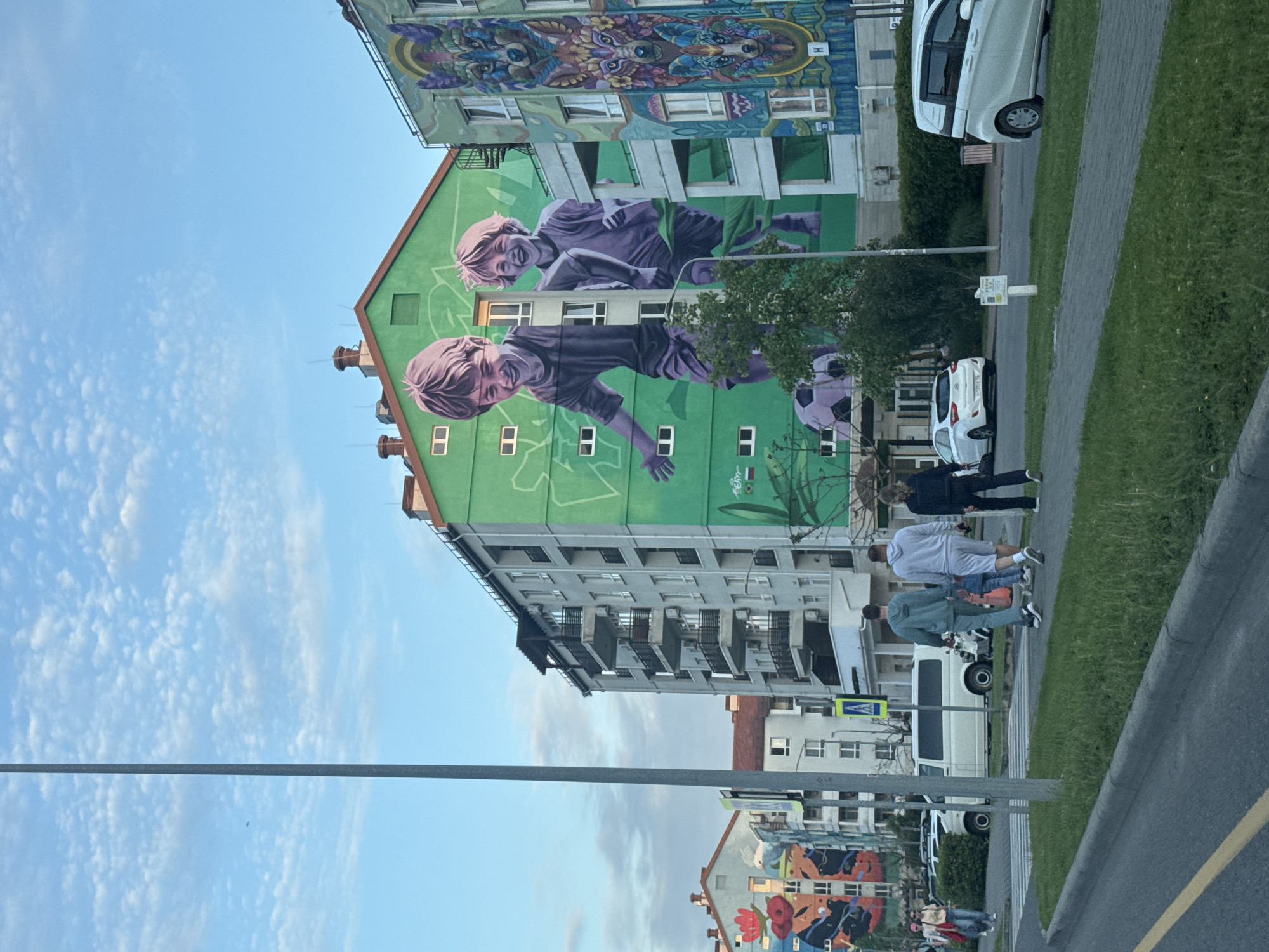 The image depicts a street scene in the city of Sirius, Russia. A multi-story building with a large mural of two boys playing soccer dominates the view. A mural of an animal is above it. A few people are walking on the sidewalk along the street. Several vehicles, including cars and vans, are parked along the side of the road. Grass and trees are also visible along the sidewalk. The sky is blue with some clouds. A street lamp is on the left side of the image.