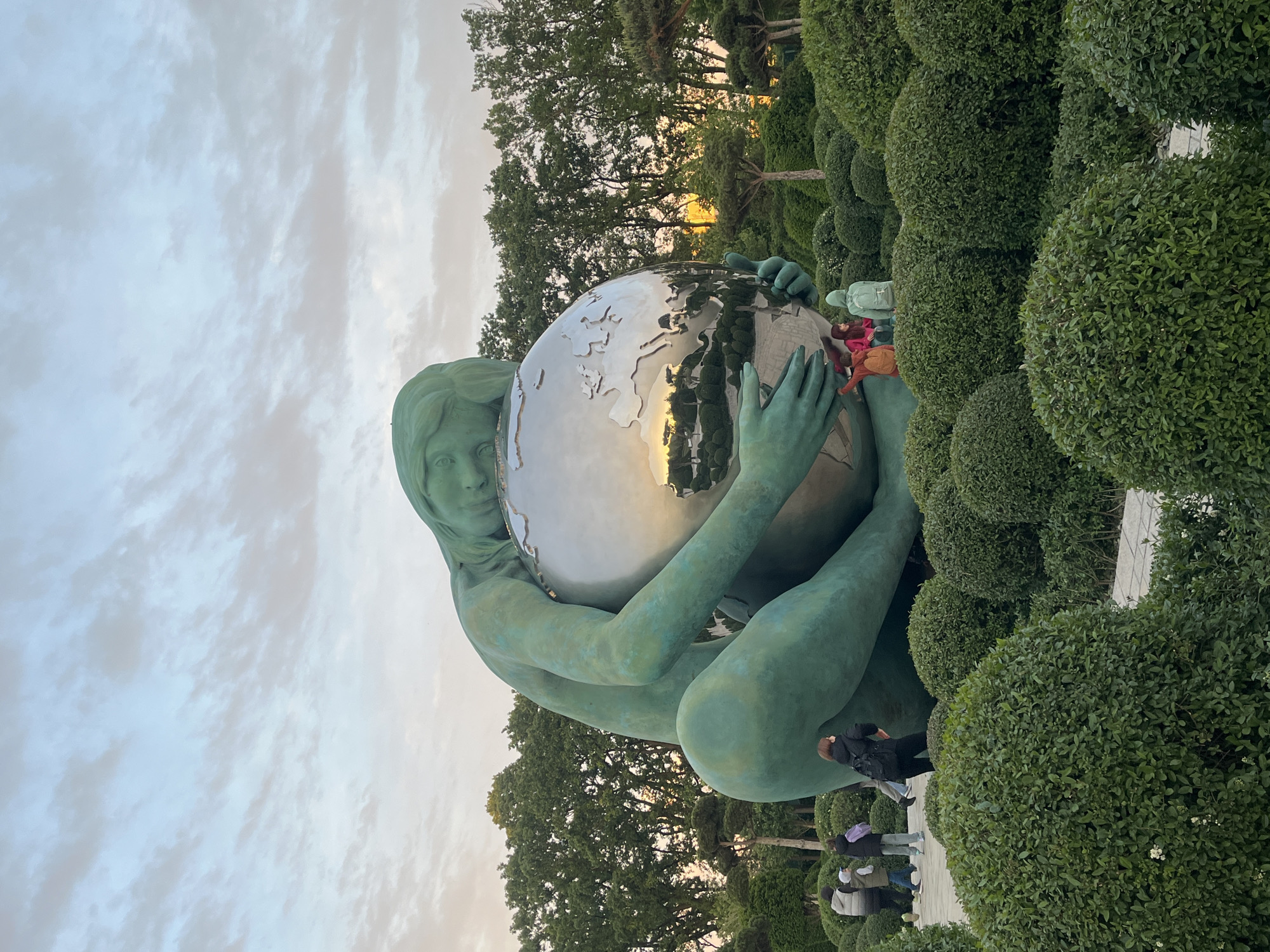 The image features a large, greenish-bronze statue of a female figure, often referred to as "Mother Earth" or "Pregnant Earth," cradling a highly reflective, silver-toned globe. The statue is situated outdoors in a public park, specifically Galitsky Park in Krasnodar, Russia.
The female figure is depicted with a serene expression, her body and hands enveloping the polished globe. The globe's surface brilliantly reflects the surrounding environment, including the cloudy sky above, green trees, and the warm, golden light suggestive of late afternoon or early evening.
The statue is surrounded by meticulously sculpted, rounded green bushes and larger, mature trees in the background. Several people are visible in the mid-ground and background, walking on paved paths or observing the artwork, providing a sense of scale to the massive sculpture. The sky is partially cloudy, casting a soft, diffused light over the scene. The overall setting is a well-maintained, serene park atmosphere. No visible text is present in the image.