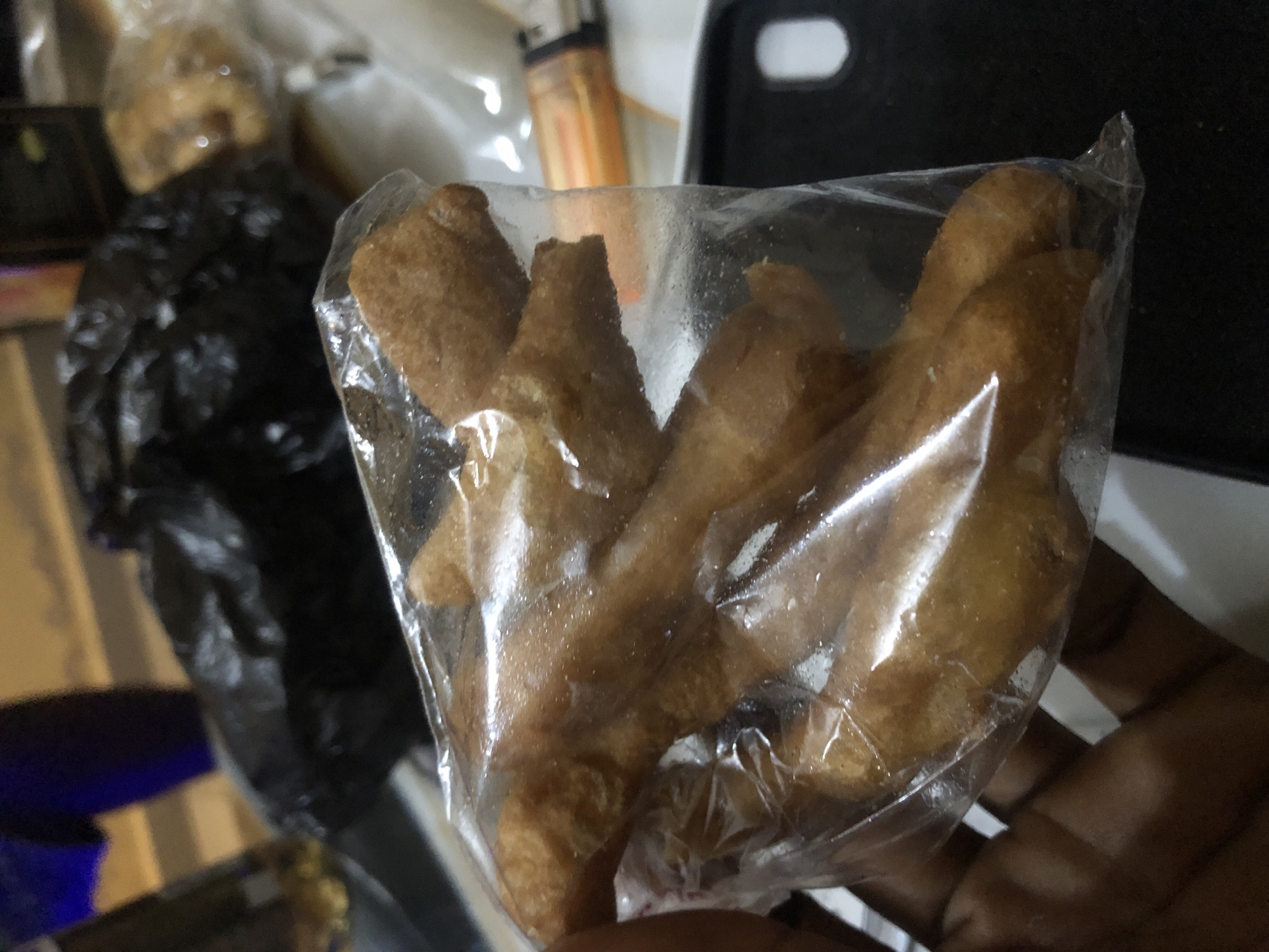 The image shows a person's hand holding a clear plastic bag filled with several golden-brown fried snacks. The snacks appear to be elongated and have a textured surface. The background is blurred but suggests an indoor setting with various objects, including a black plastic bag and what looks like a lighter. There is no visible information in the image that indicates the location is Kabong, Nigeria.
