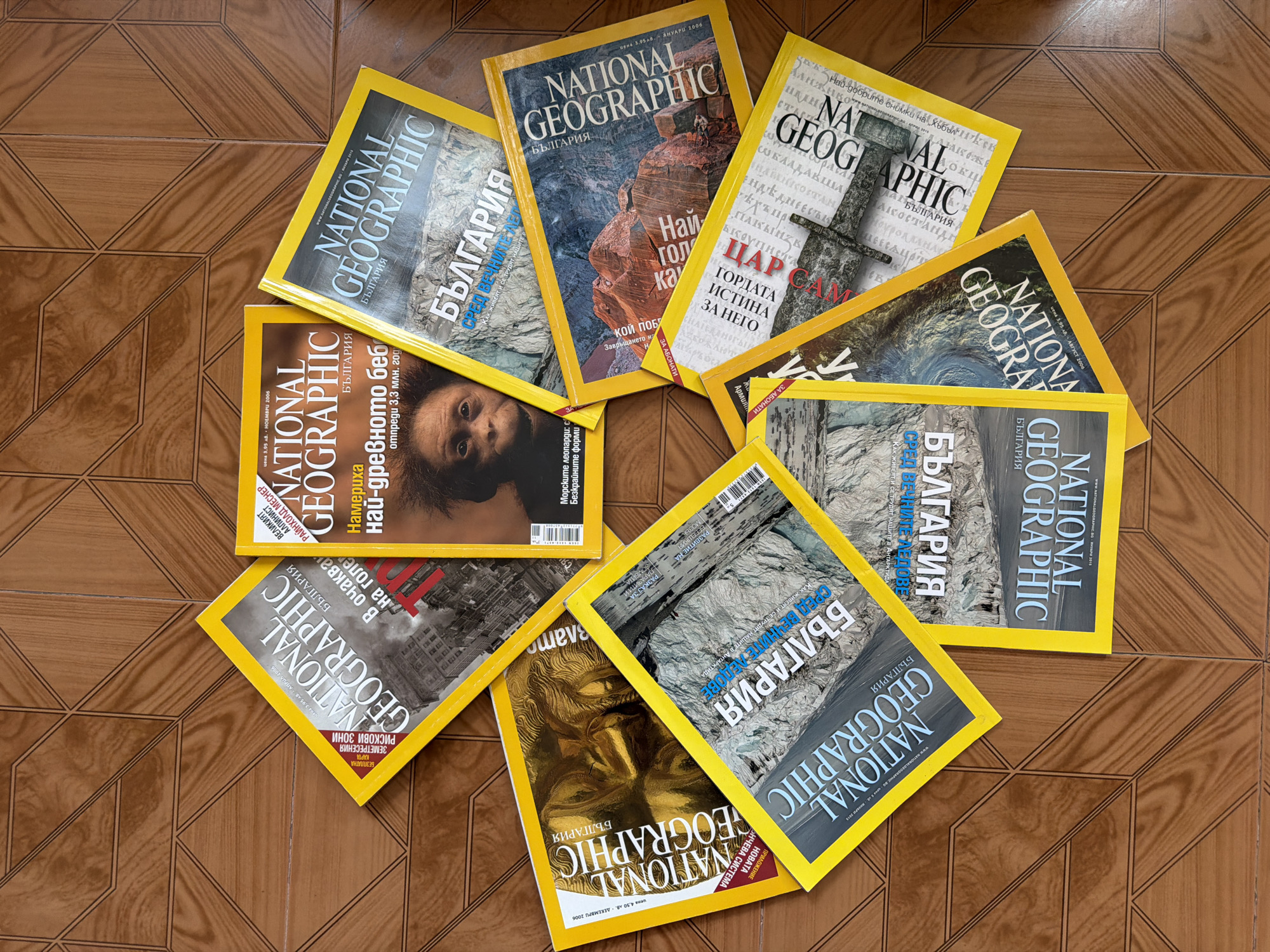 The image displays a collection of "National Geographic - Bulgaria" magazines arranged in a circular pattern on a brown, geometrically-patterned surface, possibly a tiled floor. The magazines have yellow borders with a consistent title design. The covers feature various images and titles in Bulgarian. The subjects of the magazine covers include landscapes, historical figures, human faces, and architectural elements. The arrangement is somewhat disorganized and appears to be on a floor, suggesting a casual setting. The location is not explicitly discernable within the context of the image or associated provided OCR.