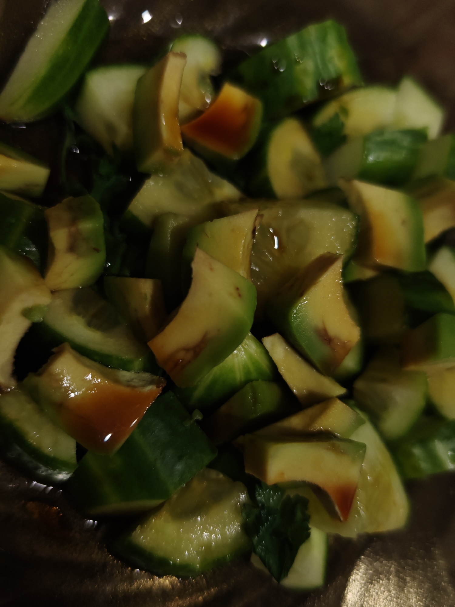 A close-up, overhead shot shows a salad made of chopped avocado and cucumber in a shallow bowl. The avocado pieces are light green to yellow with some brown spots, and the cucumber pieces are bright green. Some pieces have a shiny sheen, suggesting they are dressed with oil or a vinaigrette. A few green herbs are scattered throughout the salad. The lighting is dim and the focus is shallow, creating a blurry background that hints at a reflective surface, possibly a table. The salad appears to be prepared and ready to eat.