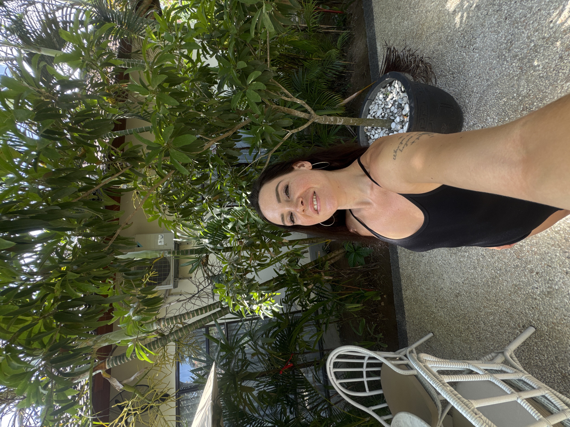 In Karangasem, Indonesia, a woman with dark hair and hoop earrings takes a selfie outdoors. She is wearing a black tank top. Behind her, lush green trees and plants surround a patio with white wicker furniture. An air conditioning unit is visible on a building in the background, along with a tiled roof. The ground is paved with small, light-colored stones.