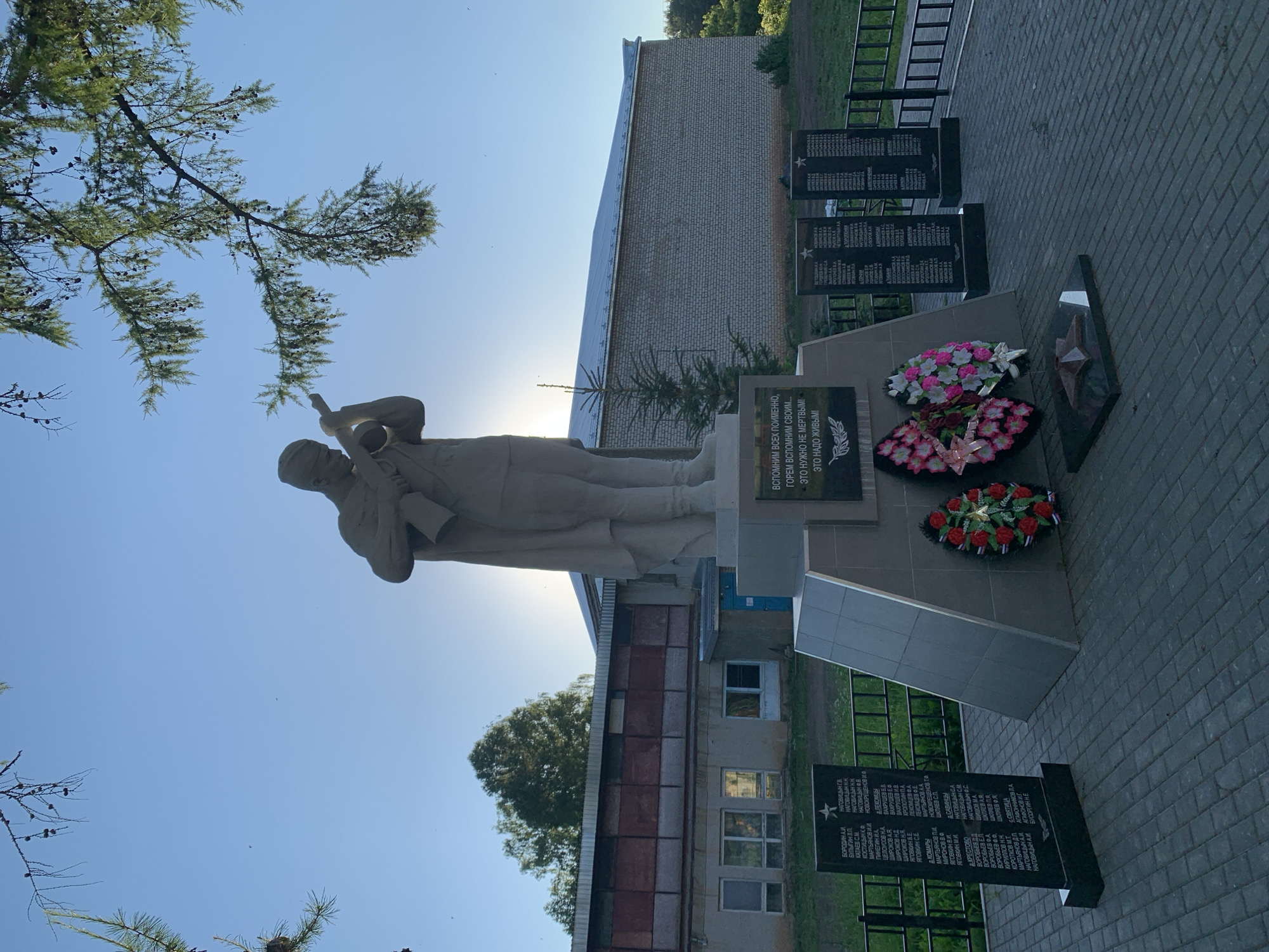 In the city of Svyatitsa, Russia, a monument stands in front of a brick building under a clear blue sky. The monument features a statue of a soldier holding a bugle. Below the statue, a central plaque reads, "LET'S REMEMBER ALL OUR FALLEN. IT IS NOT NECESSARY FOR THE DEAD - IT IS NECESSARY FOR THE LIVING."
To the left of the central plaque, there are two black granite slabs with lists of names. To the right, two more similar slabs are visible. In front of the monument, two floral wreaths are placed on the ground, one with pink and white flowers, and the other with red and pink flowers. A red star is also visible on the ground in front of the wreaths. The area around the monument is paved with light-colored square bricks. A dark metal fence encloses the monument area. The background shows a long, low-rise building with a light-colored roof. Trees are visible behind the building and to the right. A branch of a coniferous tree hangs over the top left of the image.