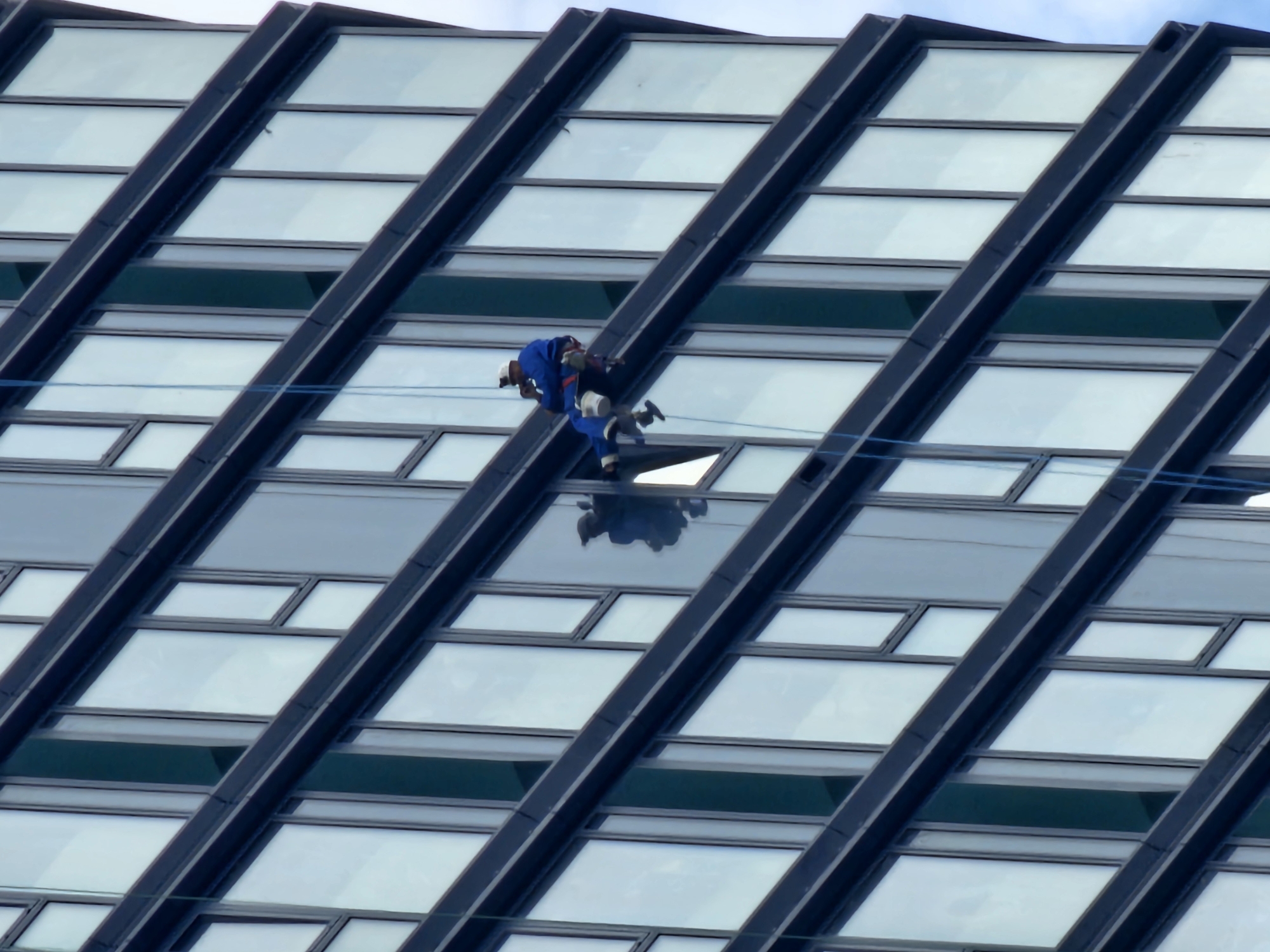 A man in blue coveralls, a white hard hat, and dark boots is cleaning the windows of a modern building in Moscow, Russia. The building features a distinctive diagonal grid pattern of dark gray or black metal beams encasing numerous rectangular windows. A set of light blue ropes secures the window cleaner. The sky is visible behind and reflected in some of the glass windows.