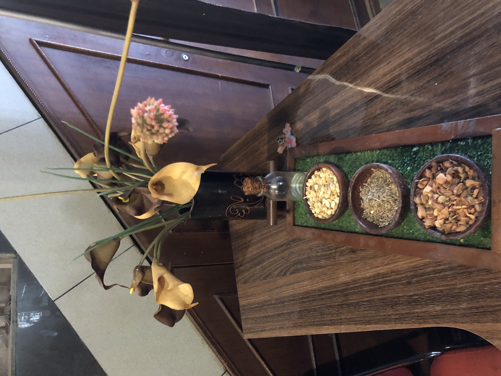 The image shows a table arrangement in what appears to be an indoor setting, possibly a restaurant or hotel in Medan, Indonesia. The table has a dark wood grain finish. On the table, there is a rectangular tray containing three small bowls filled with different dried items, possibly spices or herbs. Green artificial grass acts as a filler within the tray. Next to the tray, there is a decorative flower arrangement in a dark-colored vase. The arrangement includes artificial calla lilies and a pink pom-pom-like flower. Behind the arrangement, part of a wooden structure is visible. The floor nearby has a tile pattern.