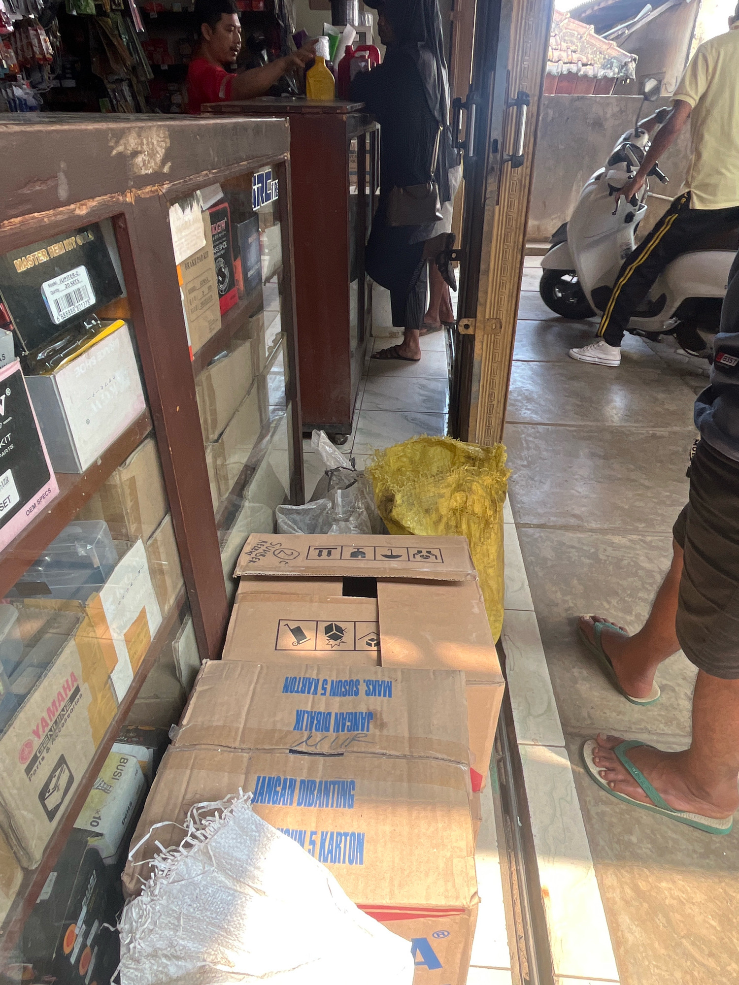The image depicts a street-level view of a shop interior and its immediate exterior in Kemuning, Indonesia. Inside the shop, a man wearing a red shirt is visible behind a counter, interacting with what appears to be store merchandise, including bottles. The shop features glass display cases filled with various goods, some of which are boxed and labeled with brand names such as "YAMAHA" and "MASTER REM KIT GOLD." Additional cardboard boxes are stacked on shelves and on the floor.
Outside the shop, a paved pathway or street is visible, with a white scooter parked. Several individuals are present. One person, wearing a yellow shirt and dark pants with a yellow stripe, is seated on the scooter. Another person, wearing dark shorts and a dark grey shirt, is standing near the entrance, their feet clad in flip-flops. A second individual, wearing dark pants and flip-flops, is also visible with their feet in the foreground. The environment appears to be an outdoor commercial area, characterized by its natural lighting and the presence of both shop interiors and exterior elements.