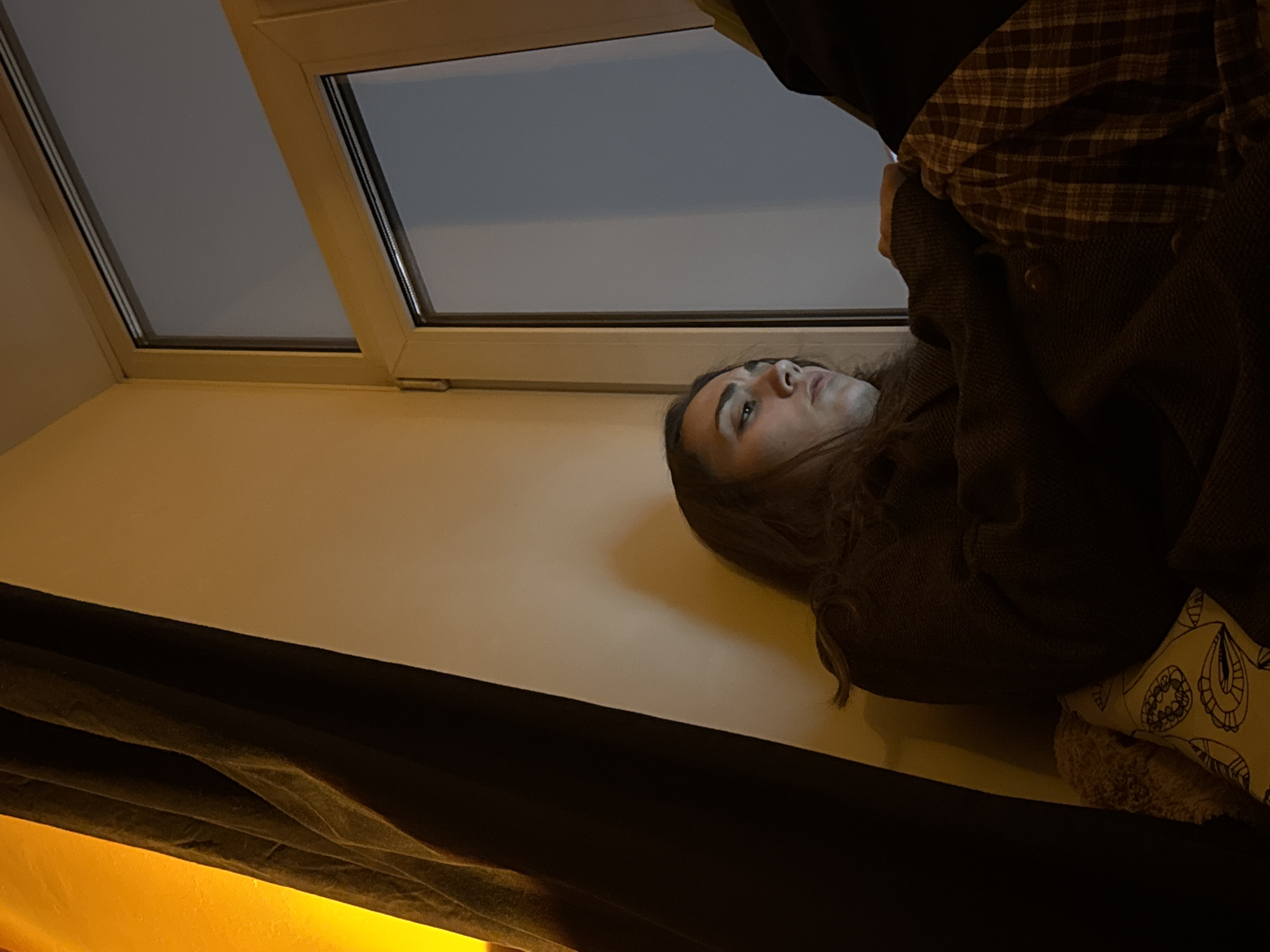The image shows a person with long brown hair lying down, possibly on a bed or couch, next to a window. The window is rectangular with a white frame, and the view outside is of a gray or overcast sky. The person is wearing a brown coat over a plaid shirt. A dark curtain drapes in the foreground. The setting is likely indoors, but the location in Moscow, Russia, cannot be determined from the image alone.