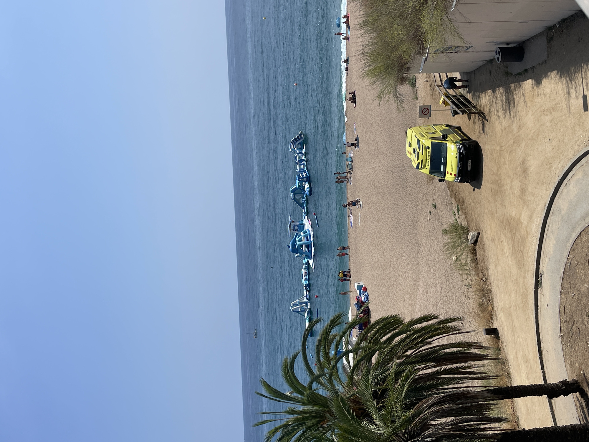 In Calella, Spain, a yellow ambulance is parked on a sandy path near the beach, its lights flashing. The ambulance is positioned facing left, with its driver's side visible. A person is bending over near a trash can to the right of the ambulance. The beach is populated with numerous people sunbathing and swimming. In the water, an inflatable water park with various blue structures is set up, with people visible on and around it. A sailboat is in the distance on the calm sea. Palm trees frame the left side of the image. The scene suggests a typical summer day at a popular beach resort.